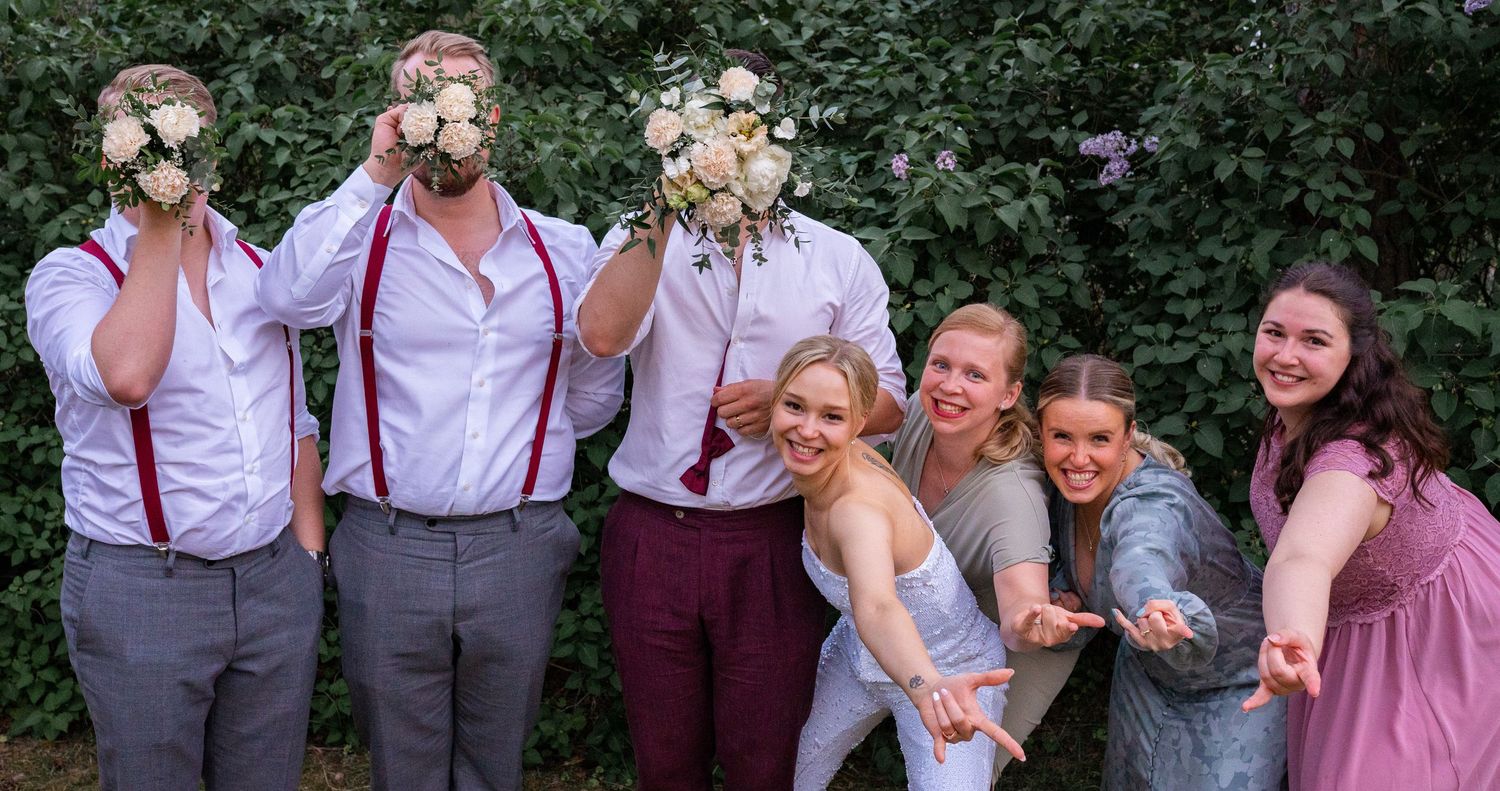 Groomsmen hiding faces with bouquets while bridesmaids pose playfully in front of lush green hedge.
