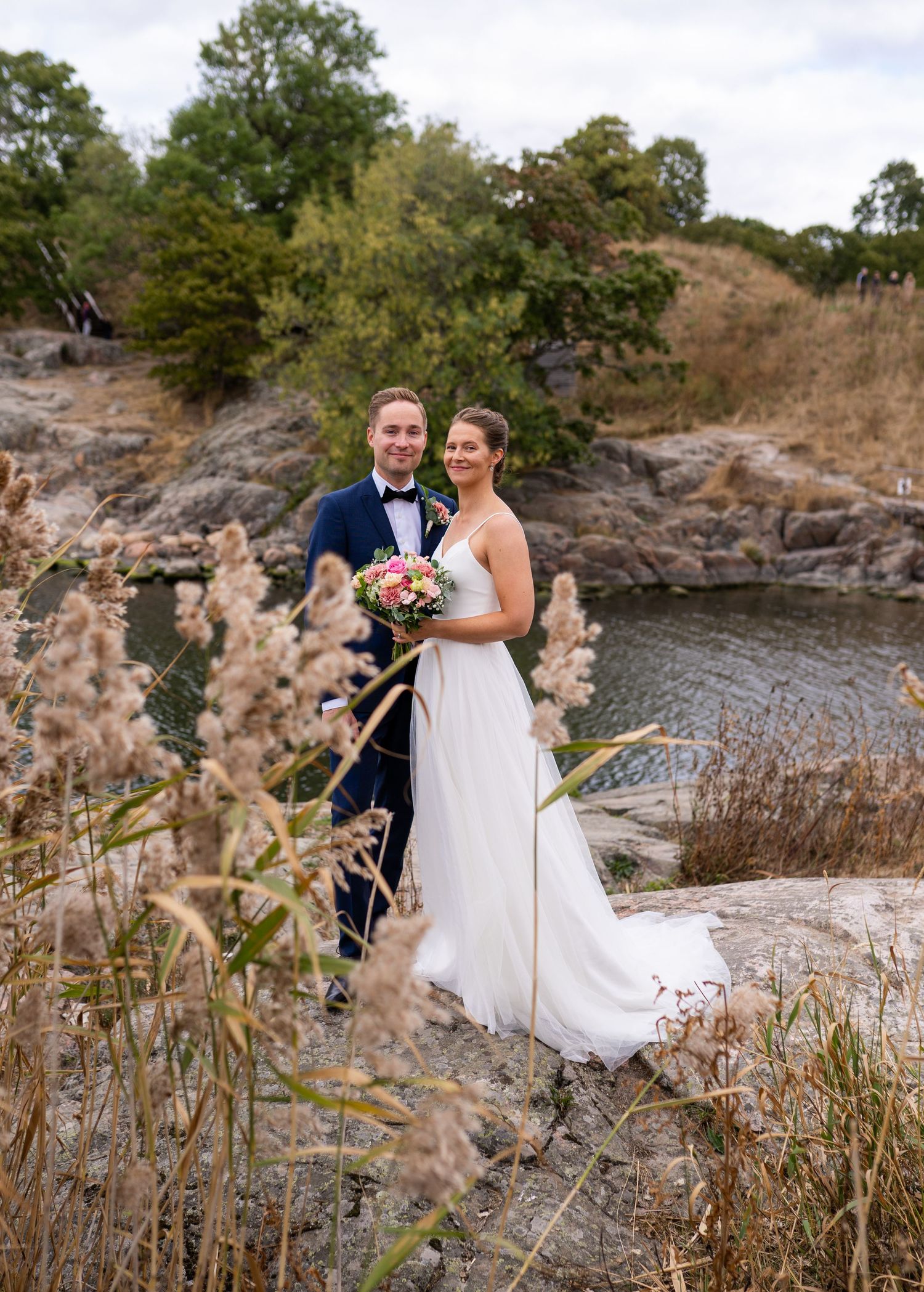 Smiling bride and groom on rocky waterside landscape, she holds pink bouquet, dried grass in foreground.