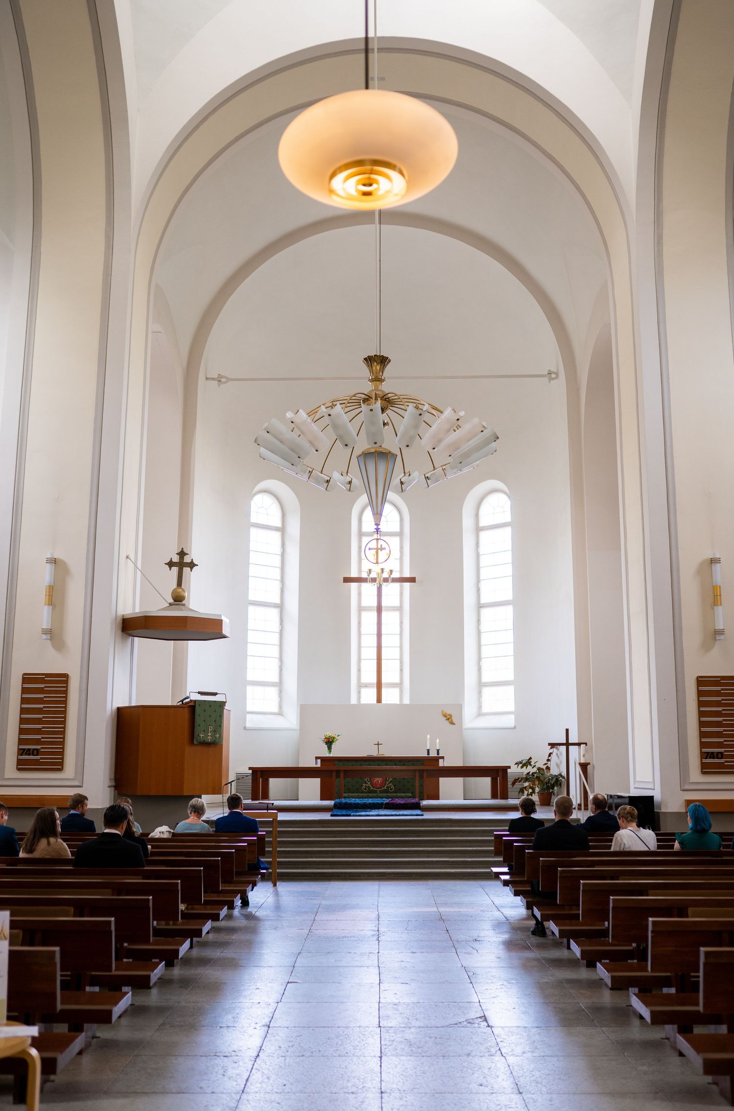 Bright white church interior with wooden pews, arched ceiling, altar cross, and chandelier during a wedding ceremony.