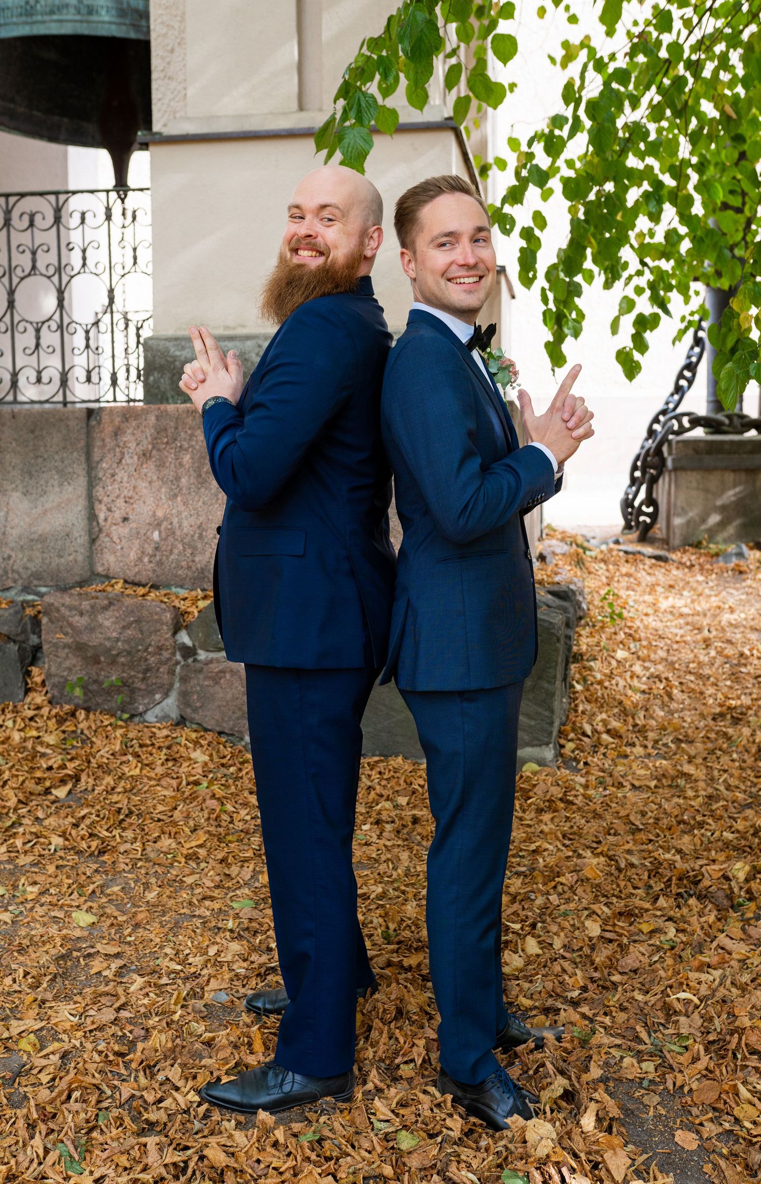 Two men in navy suits posing back-to-back on autumn leaves outside a church, pointing fingers upward playfully.