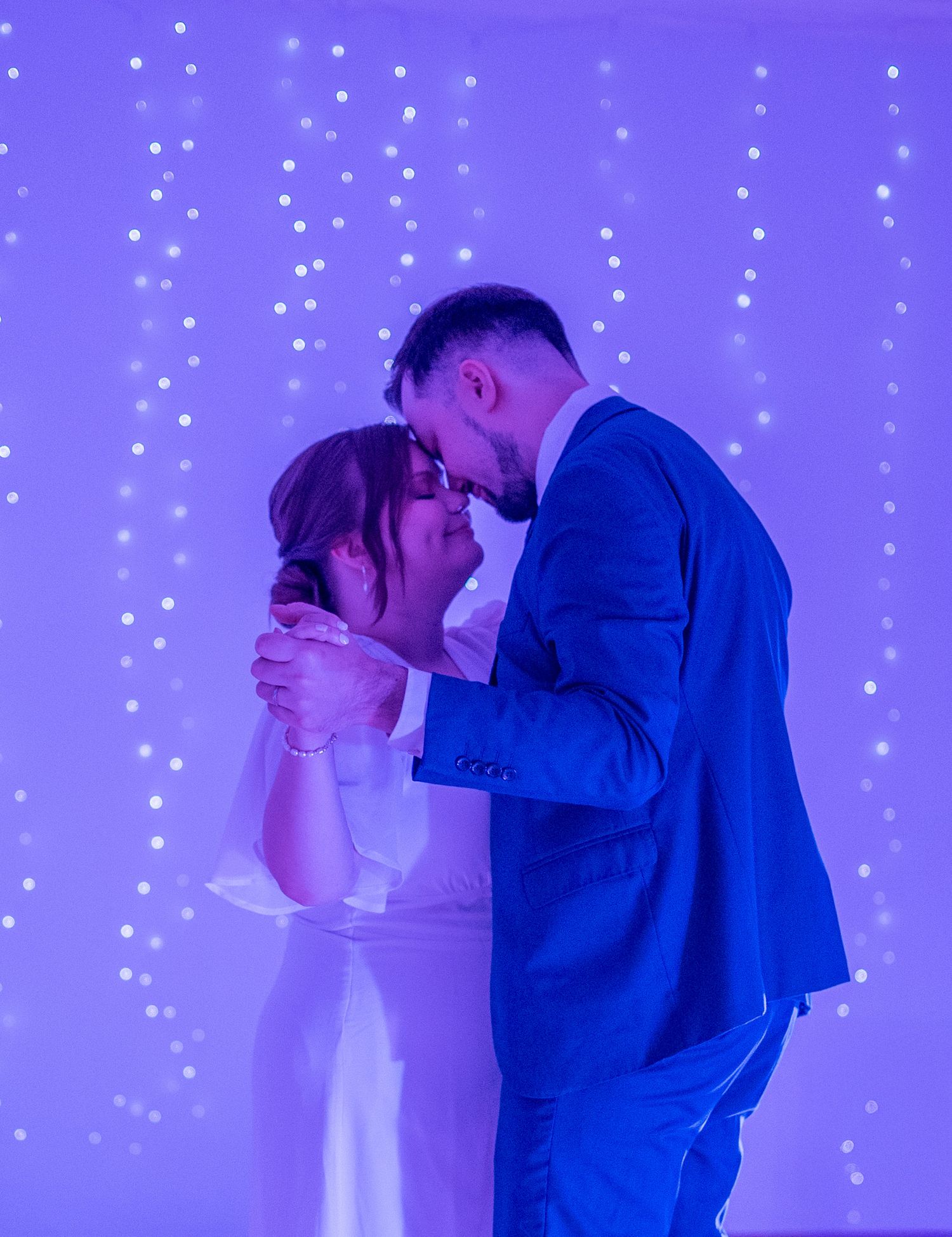 Bride and groom share intimate moment during first dance under purple fairy light backdrop.