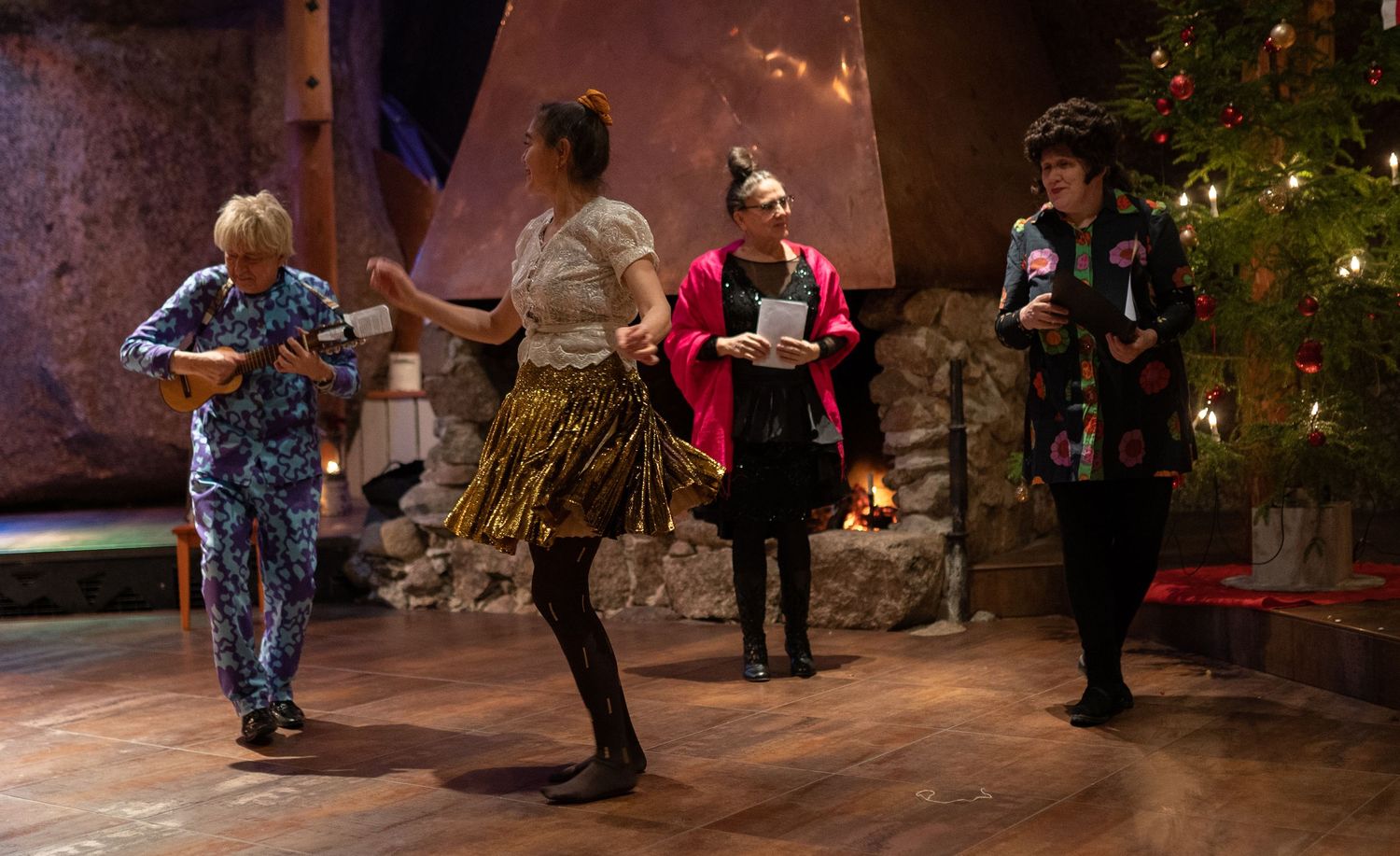 Four women dancing and celebrating near a Christmas tree in a festive indoor setting with warm lighting.