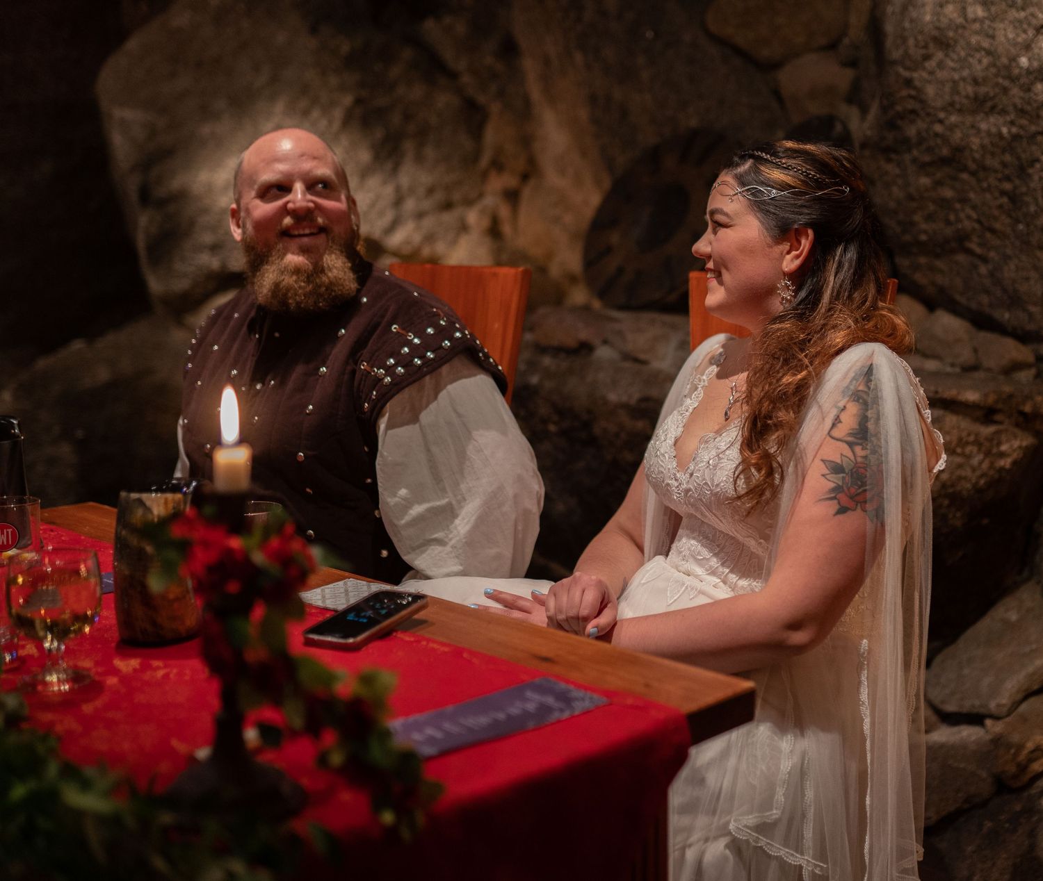 A bearded groom and tattooed bride laugh together at a candlelit medieval-themed wedding table inside a stone cave.