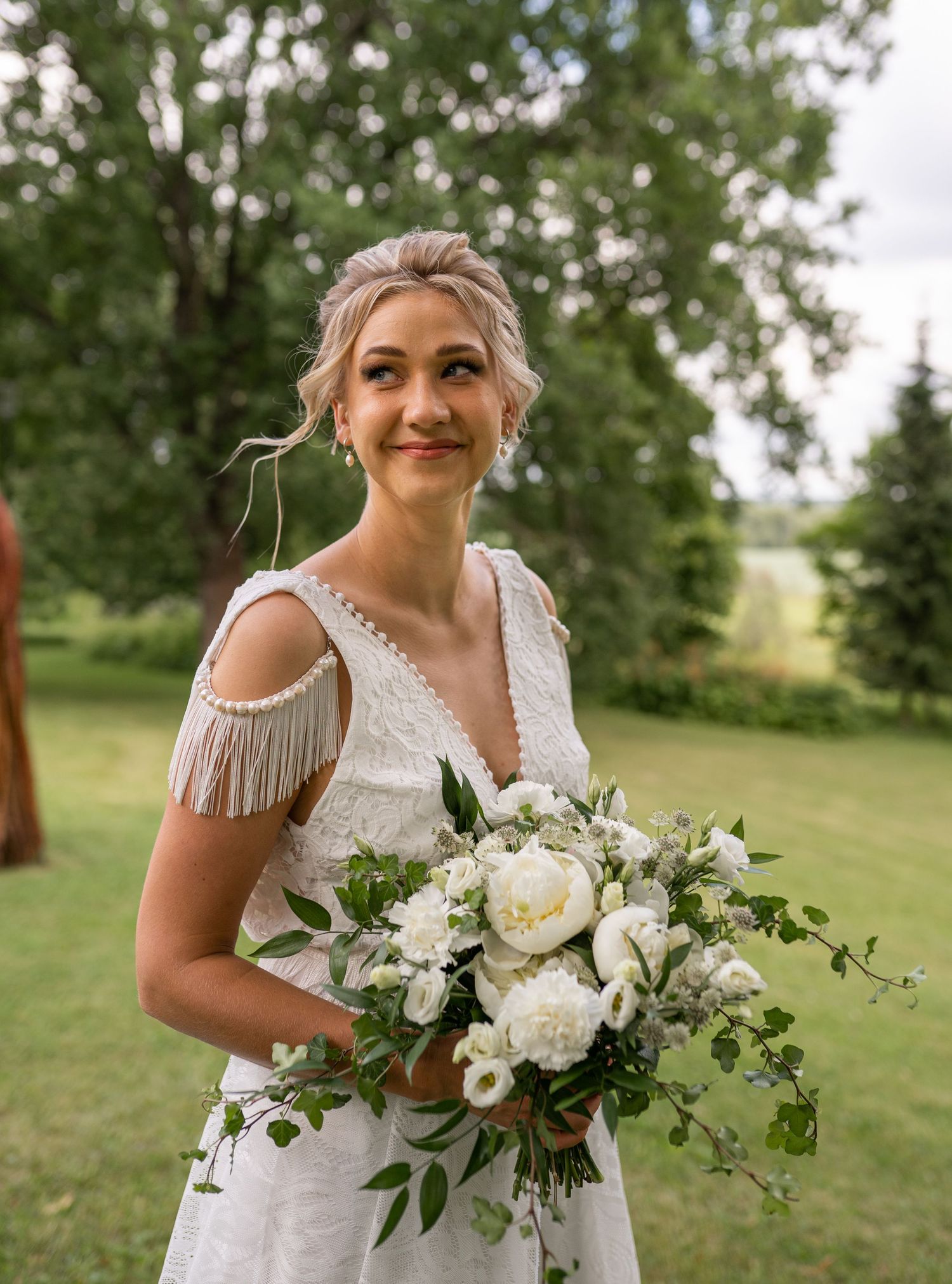 Radiant bride in lace gown with fringe shoulders holds white peony bouquet outdoors.