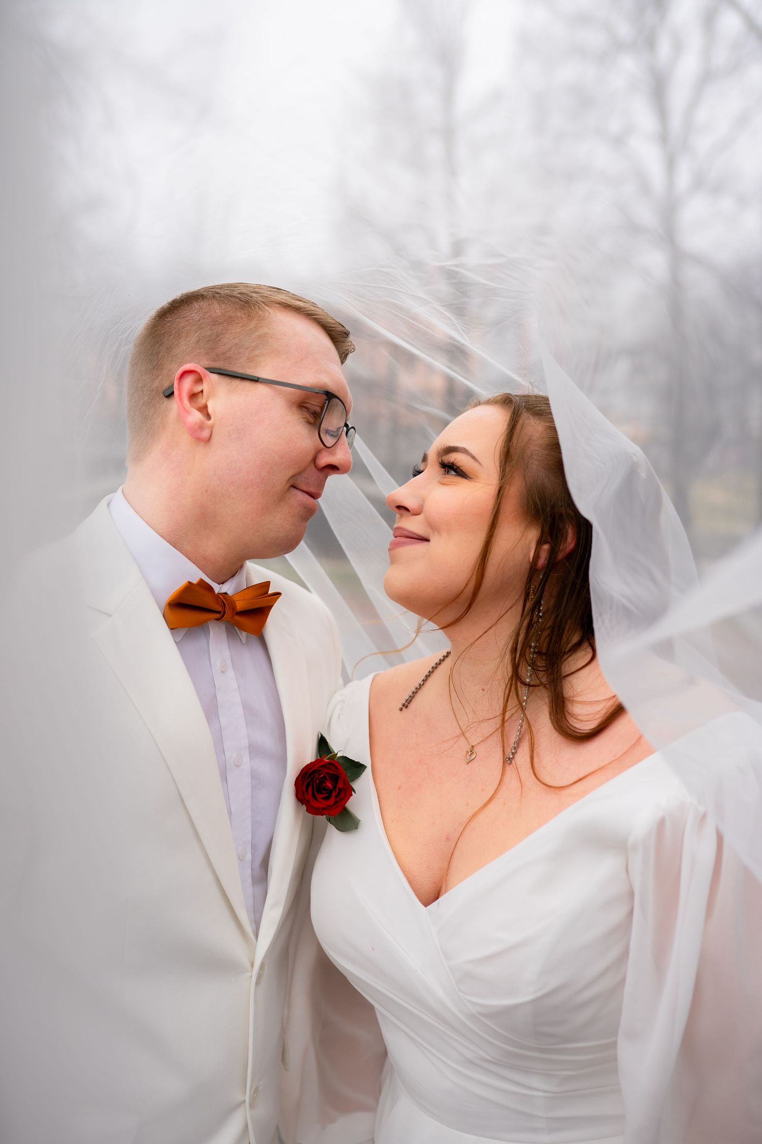 Bride and groom sharing romantic moment under flowing veil, groom wearing cream suit with orange bow tie and rose boutonniere.