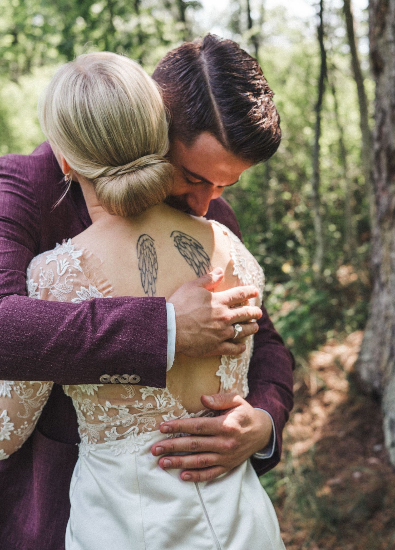 Groom in burgundy suit embraces bride with angel wings back tattoo in lace dress during outdoor wedding.