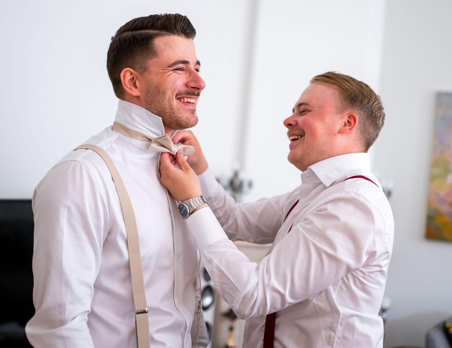 Two smiling groomsmen in white shirts with suspenders helping each other with a tie.