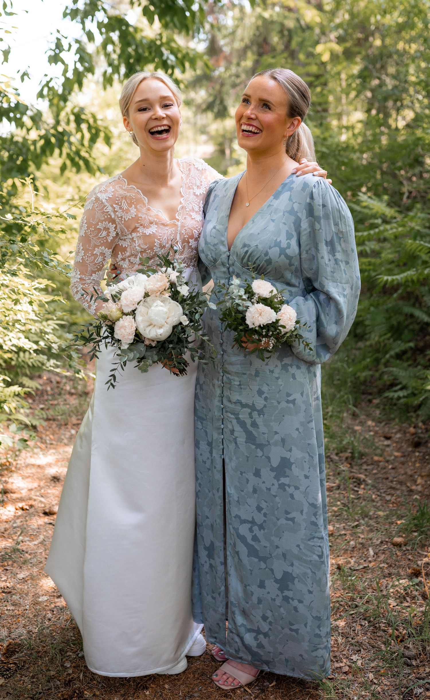 Laughing bride in lace gown and bridesmaid in blue floral dress holding bouquets.