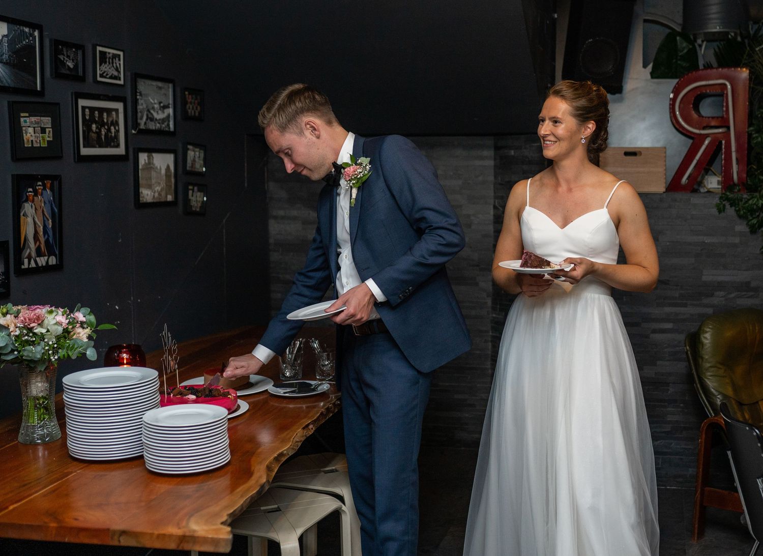 Groom serving wedding cake slices while smiling bride watches at elegant reception table.