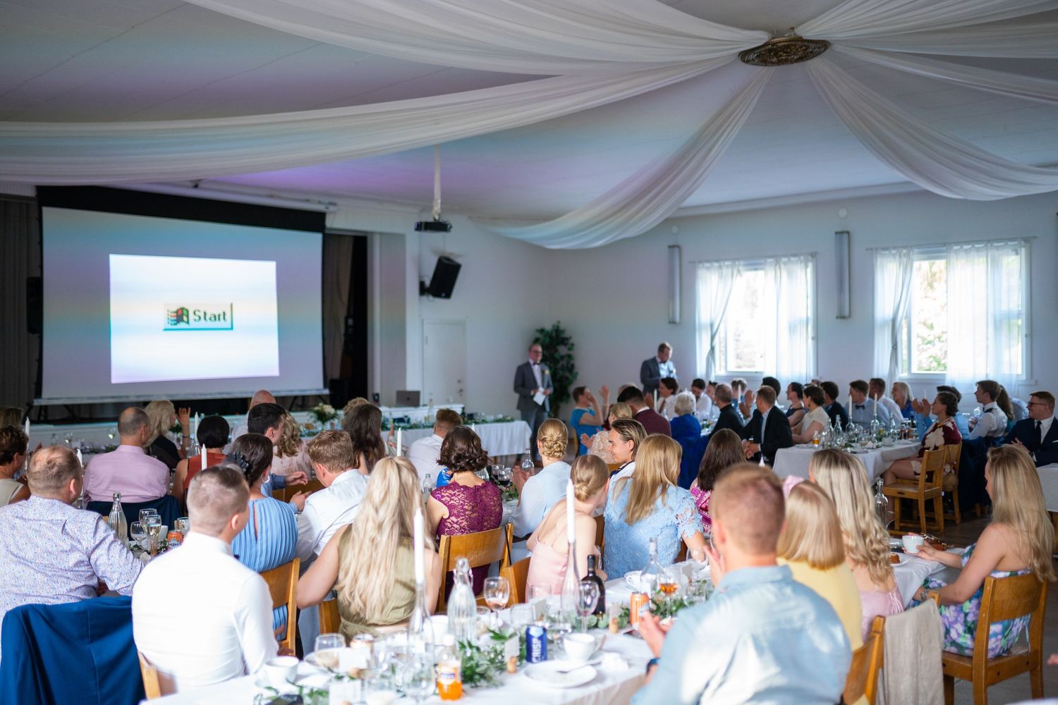 Wedding reception guests seated at decorated tables watching a presentation on a large screen.