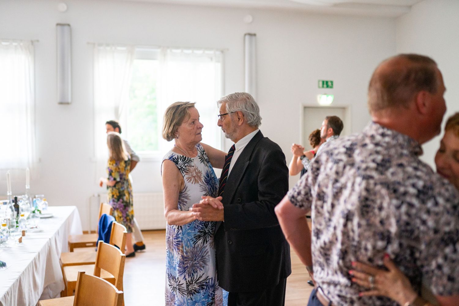 Elegant couple dancing at wedding reception in bright white hall with decorated tables nearby.