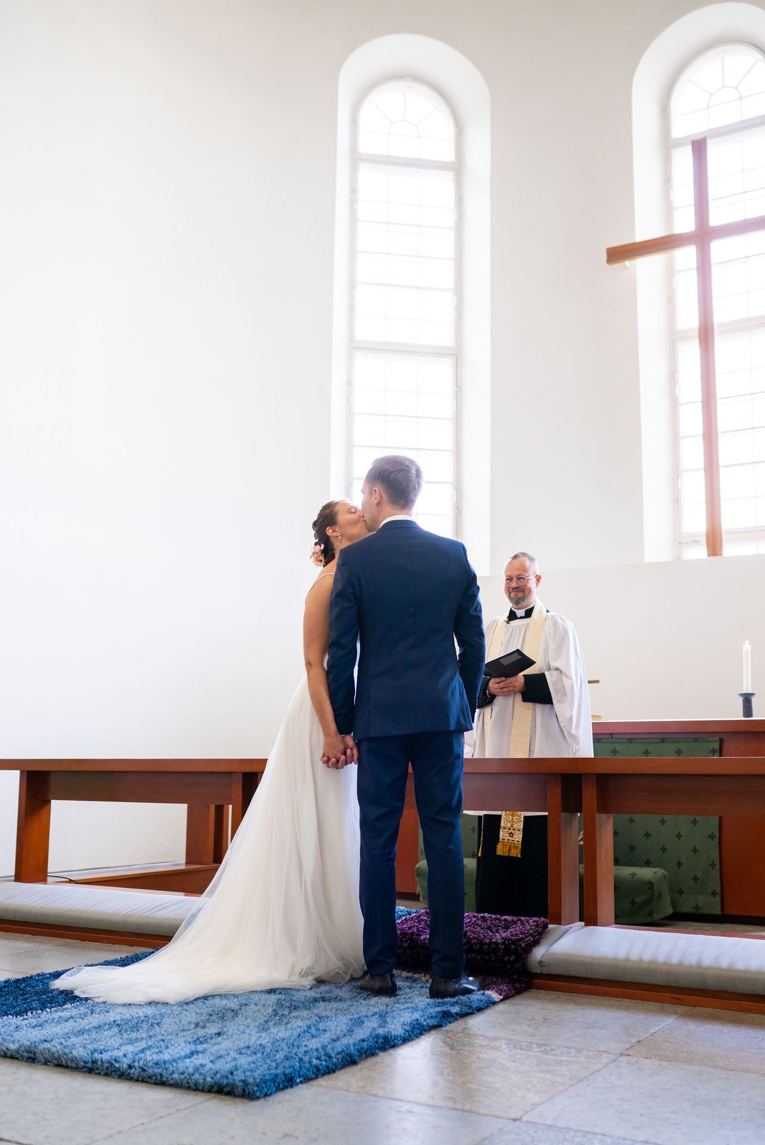 Newlyweds sharing first kiss at church altar with smiling officiant behind them.