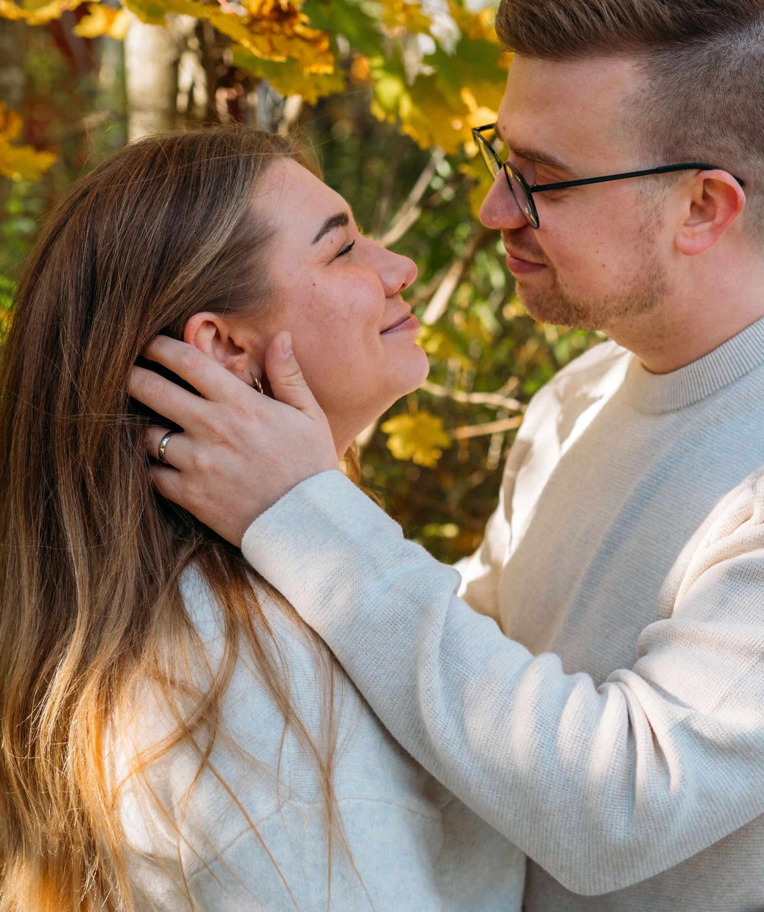 A couple shares a tender moment outdoors in autumn, man gently cupping womans face as golden leaves frame them.