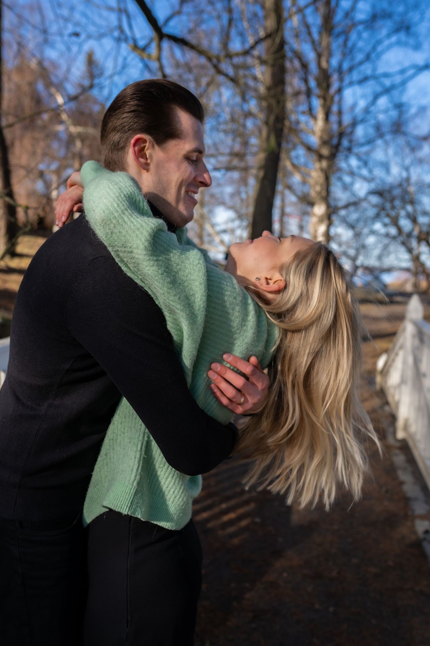 A smiling couple embraces outdoors in a park during winter, with bare trees and blue sky in the background.