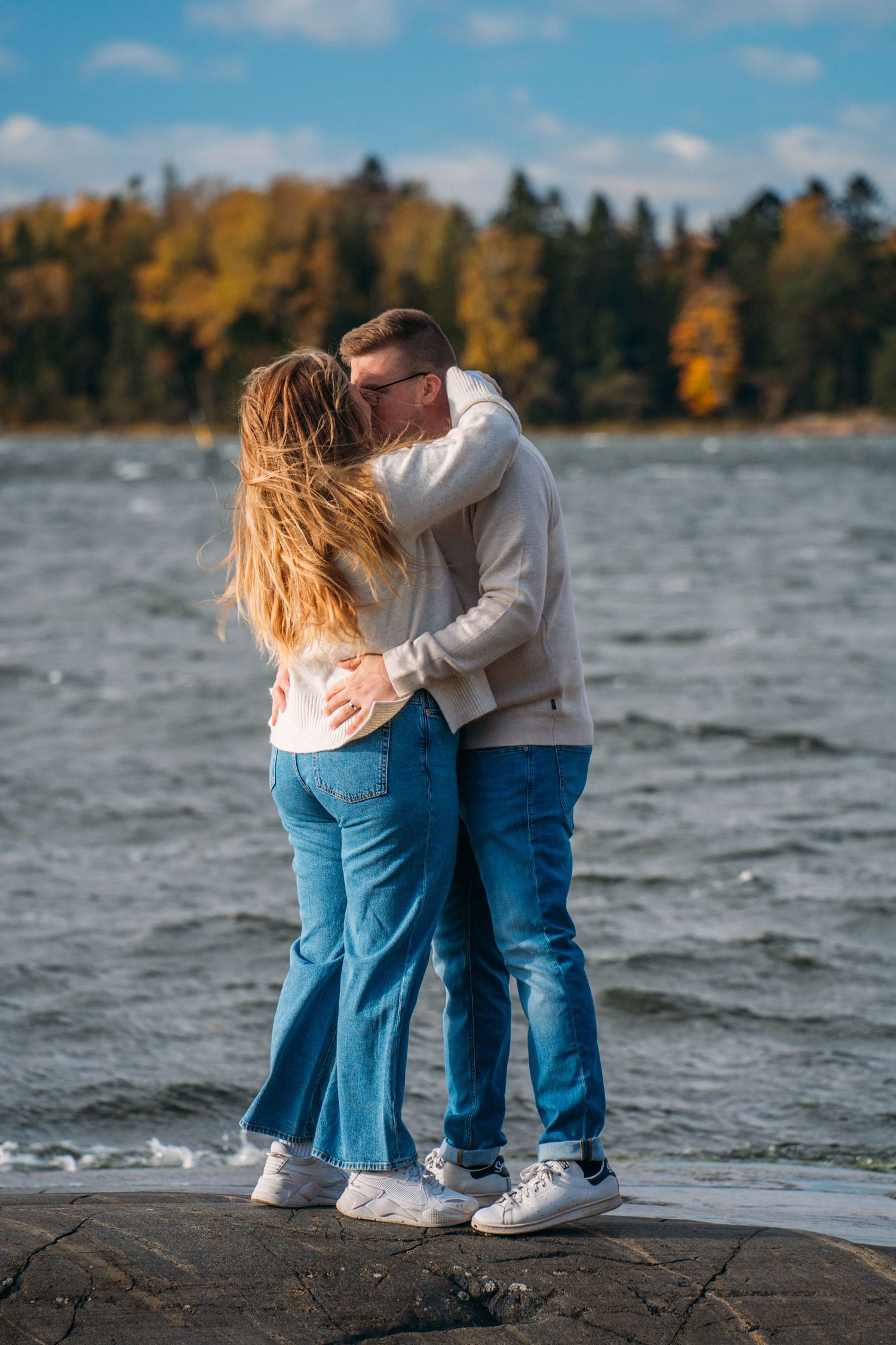 A couple sharing a romantic kiss while standing on rocks by a lake with autumn trees in the background.