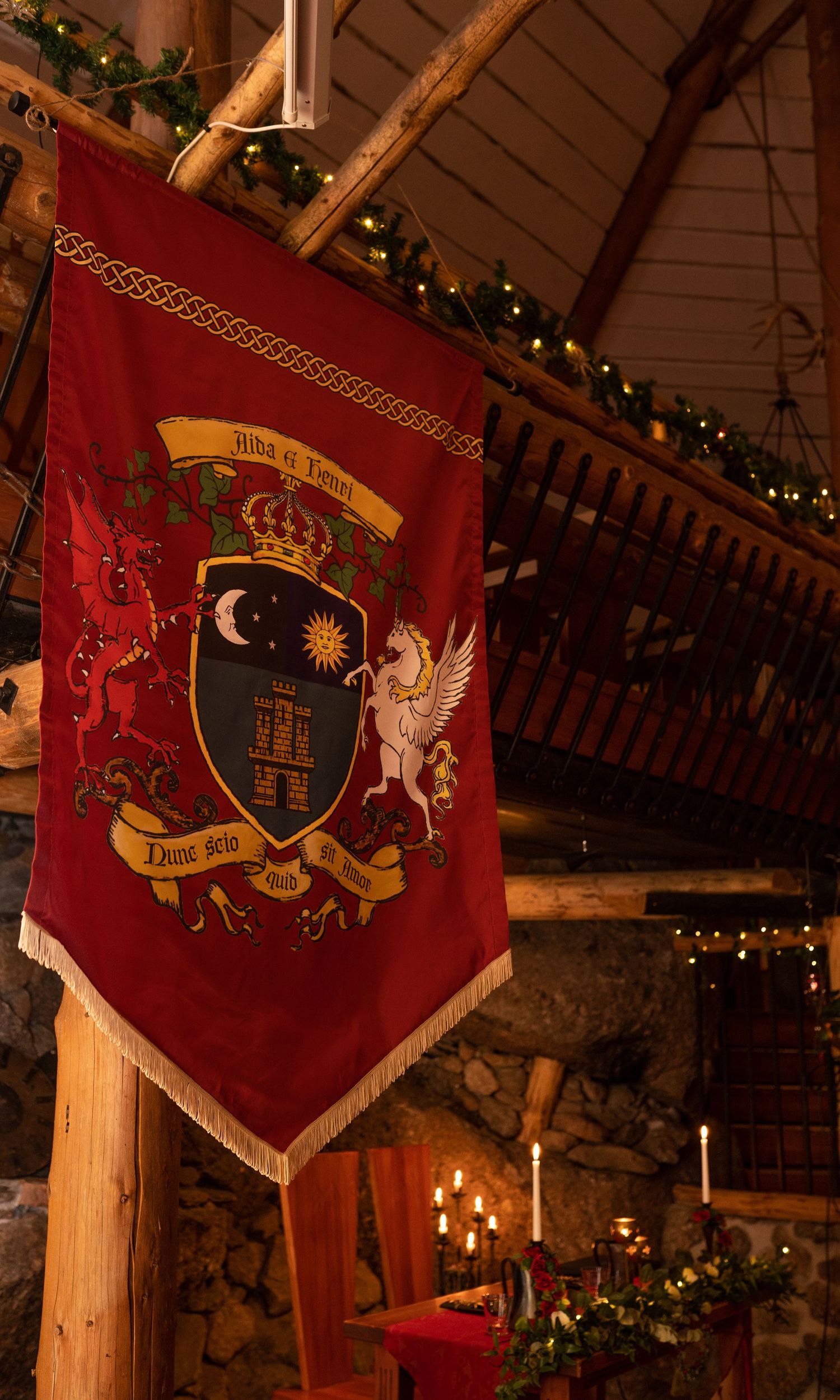 Red heraldic banner with coat of arms featuring unicorn, crown, moon and sun, hanging in decorated medieval hall.