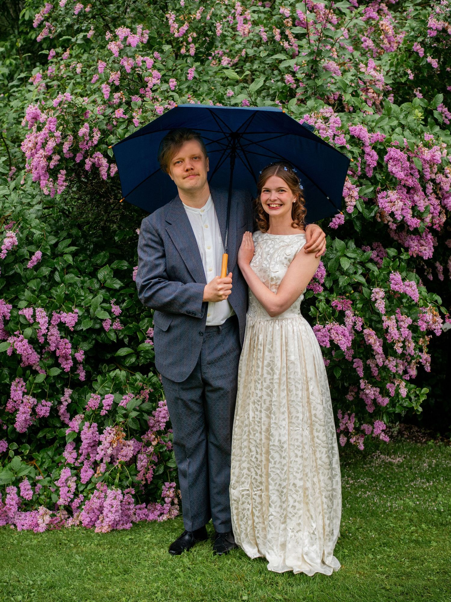 Wedding couple poses under a navy umbrella in front of blooming pink flowers in the rain.