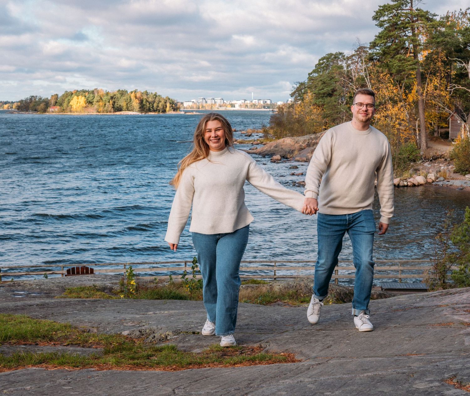 Couple holding hands walking along rocky shoreline with scenic blue water and autumn trees in background.