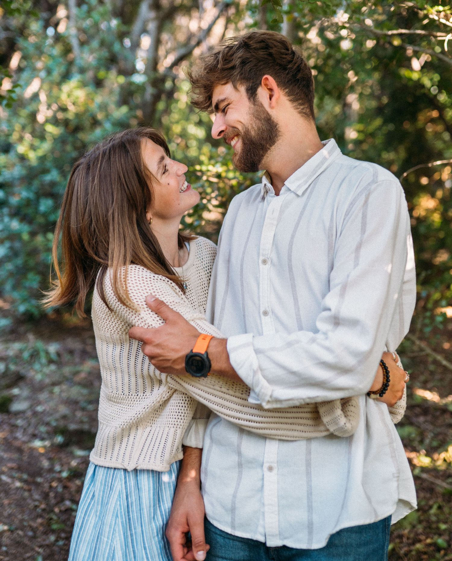 Smiling couple embracing and looking at each other on a sunny woodland trail outdoors.