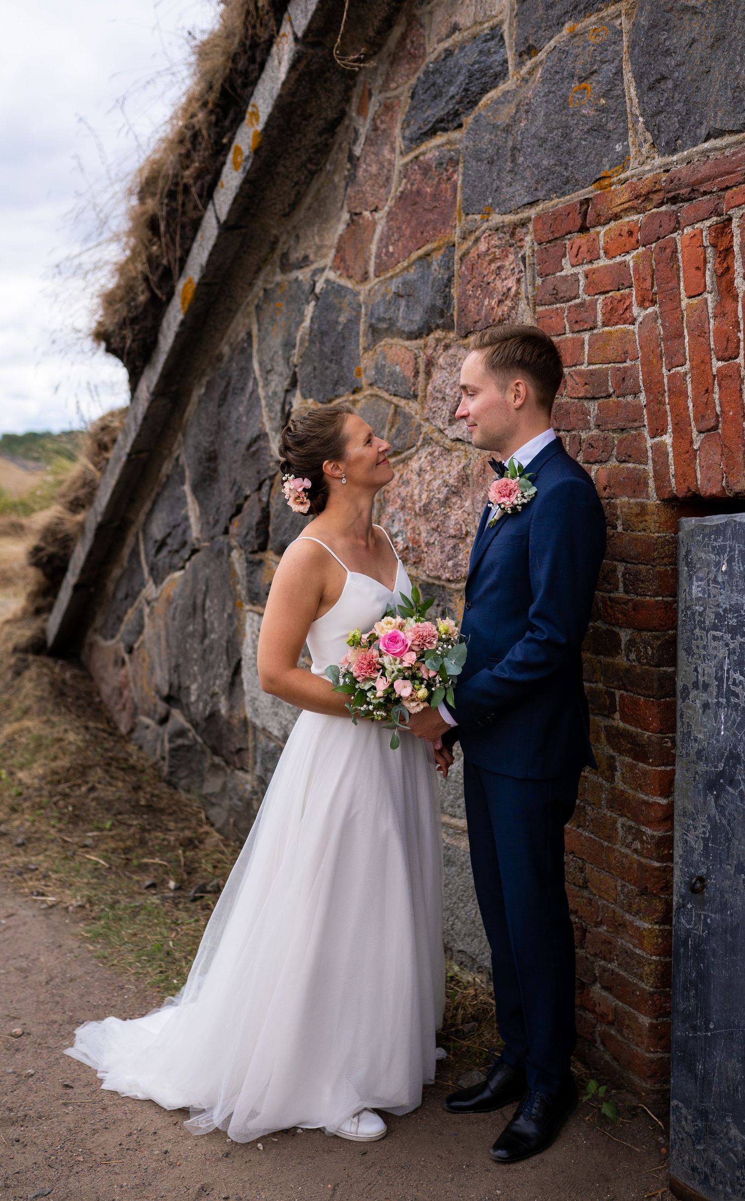 Bride in white gown and groom in navy suit facing each other against an old stone brick wall.