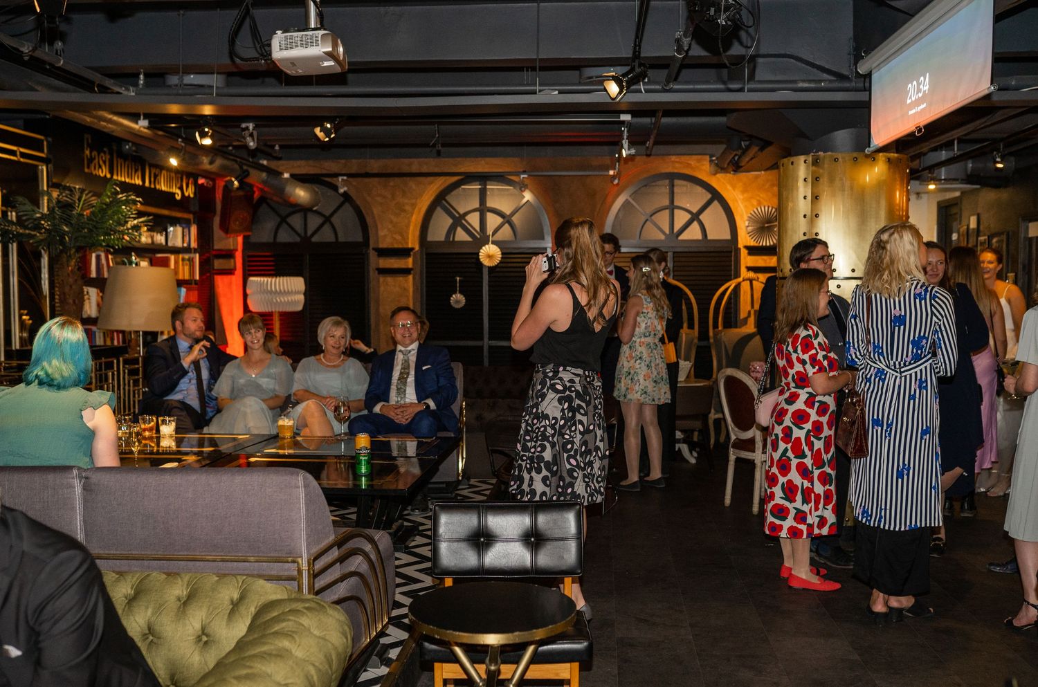 Wedding reception guests mingle in stylish venue as woman speaks to seated crowd in dark-walled room.