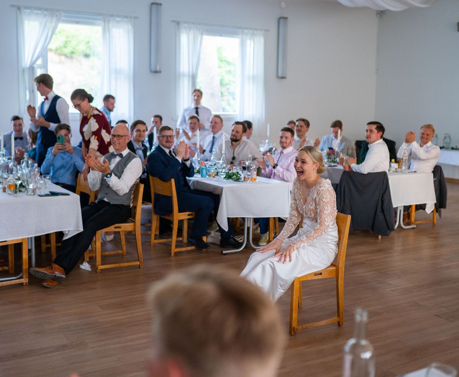 Wedding party seated at decorated reception tables, bride in lace gown laughing with guests.