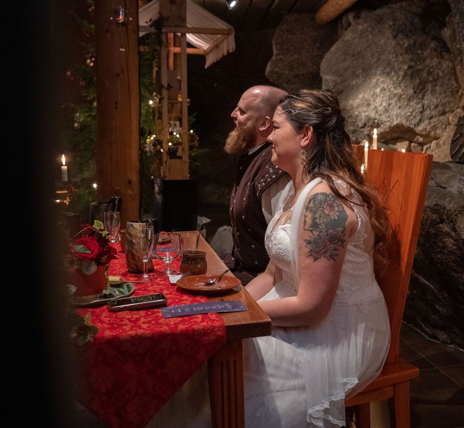Bride and groom in wedding attire seated at candlelit table with red tablecloth in rustic stone venue.