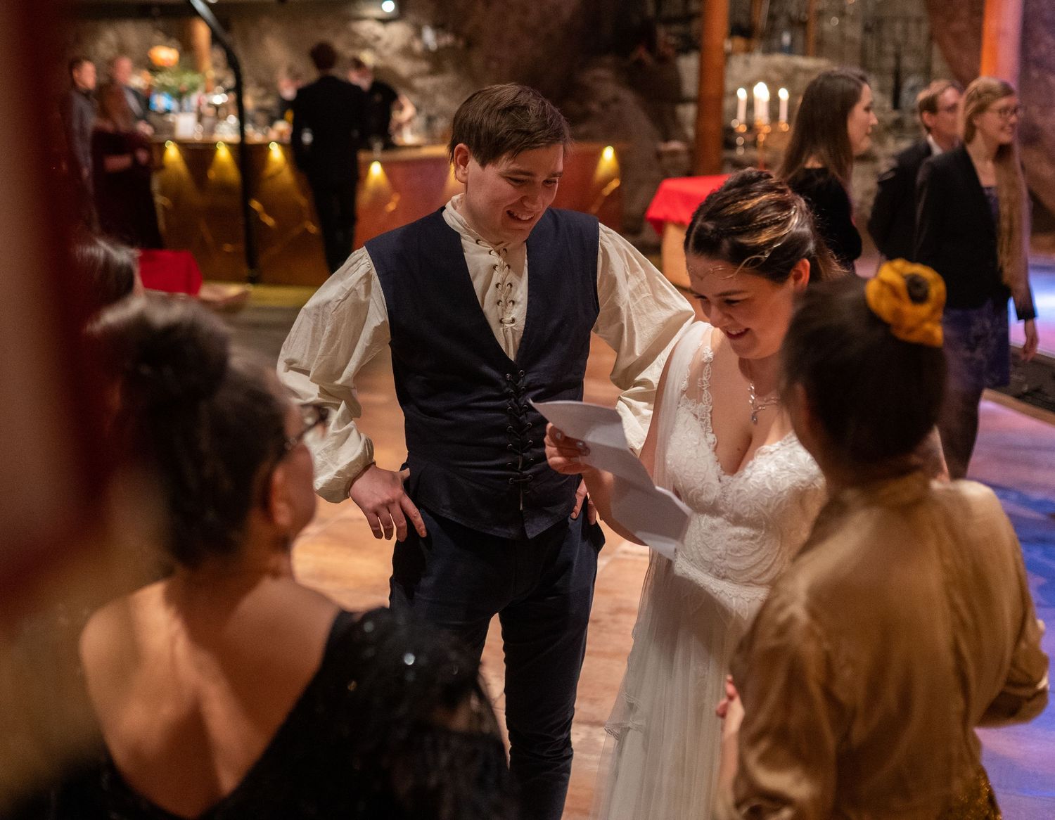 Bride and groom share a joyful moment reading vows at a rustic wedding reception with guests nearby.
