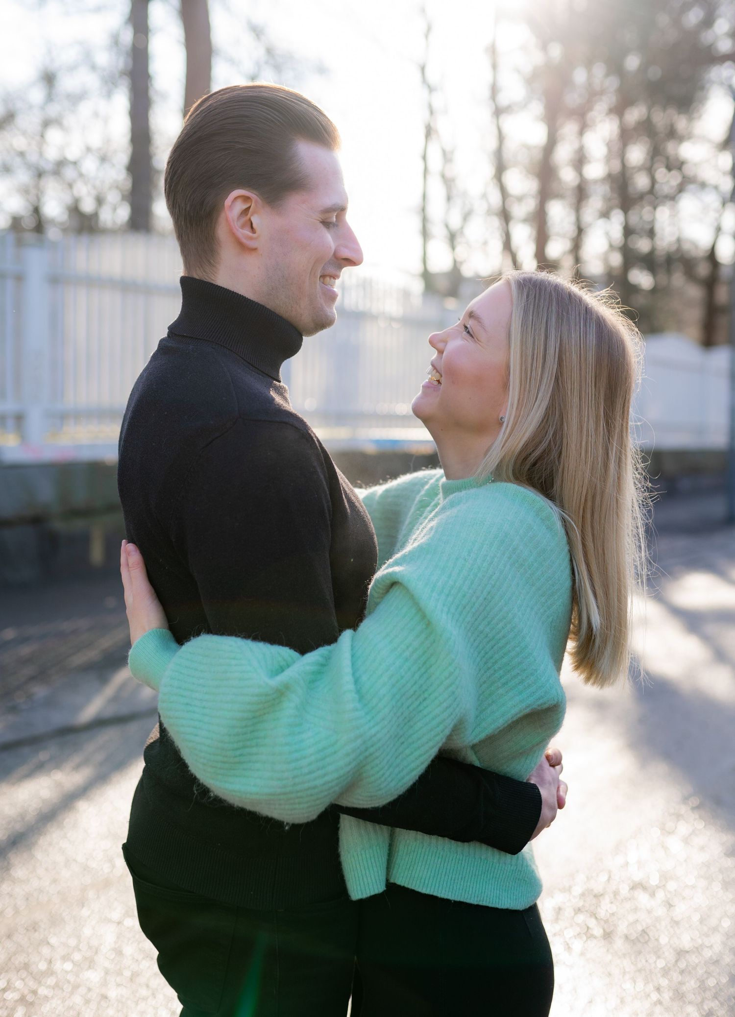 A couple embracing outdoors in golden sunlight, woman in mint green sweater gazing up at smiling man in black turtleneck.