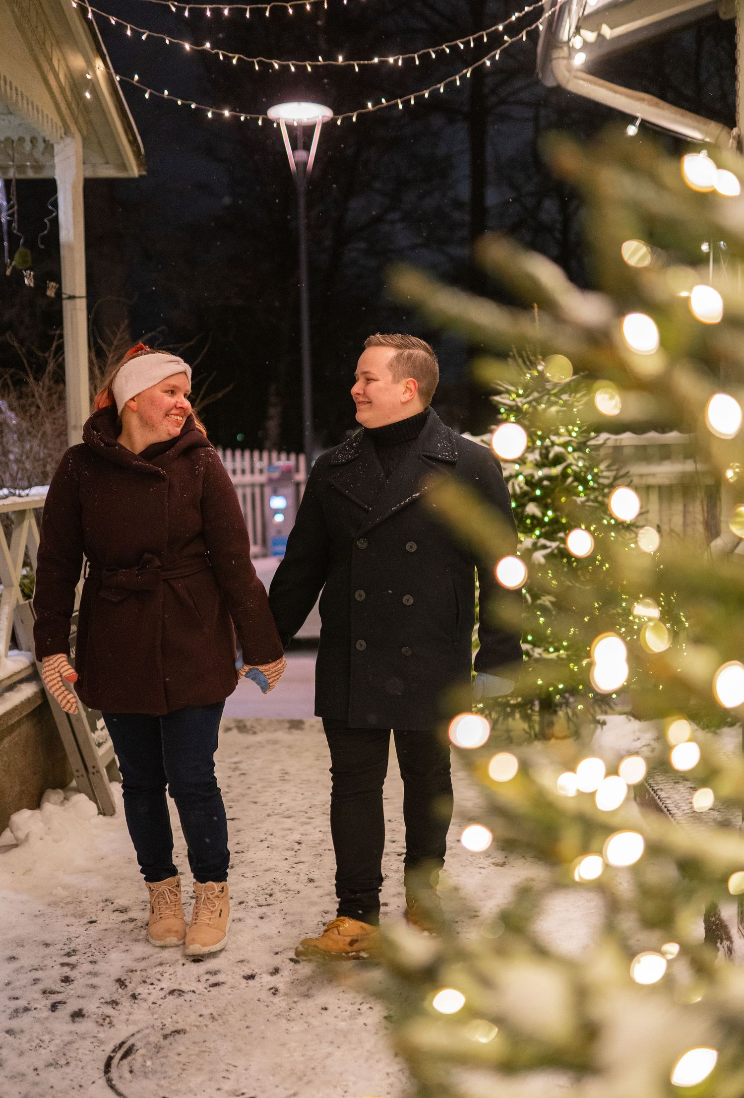 Couple holding hands walking past a decorated Christmas tree and string lights in a snowy winter evening setting.