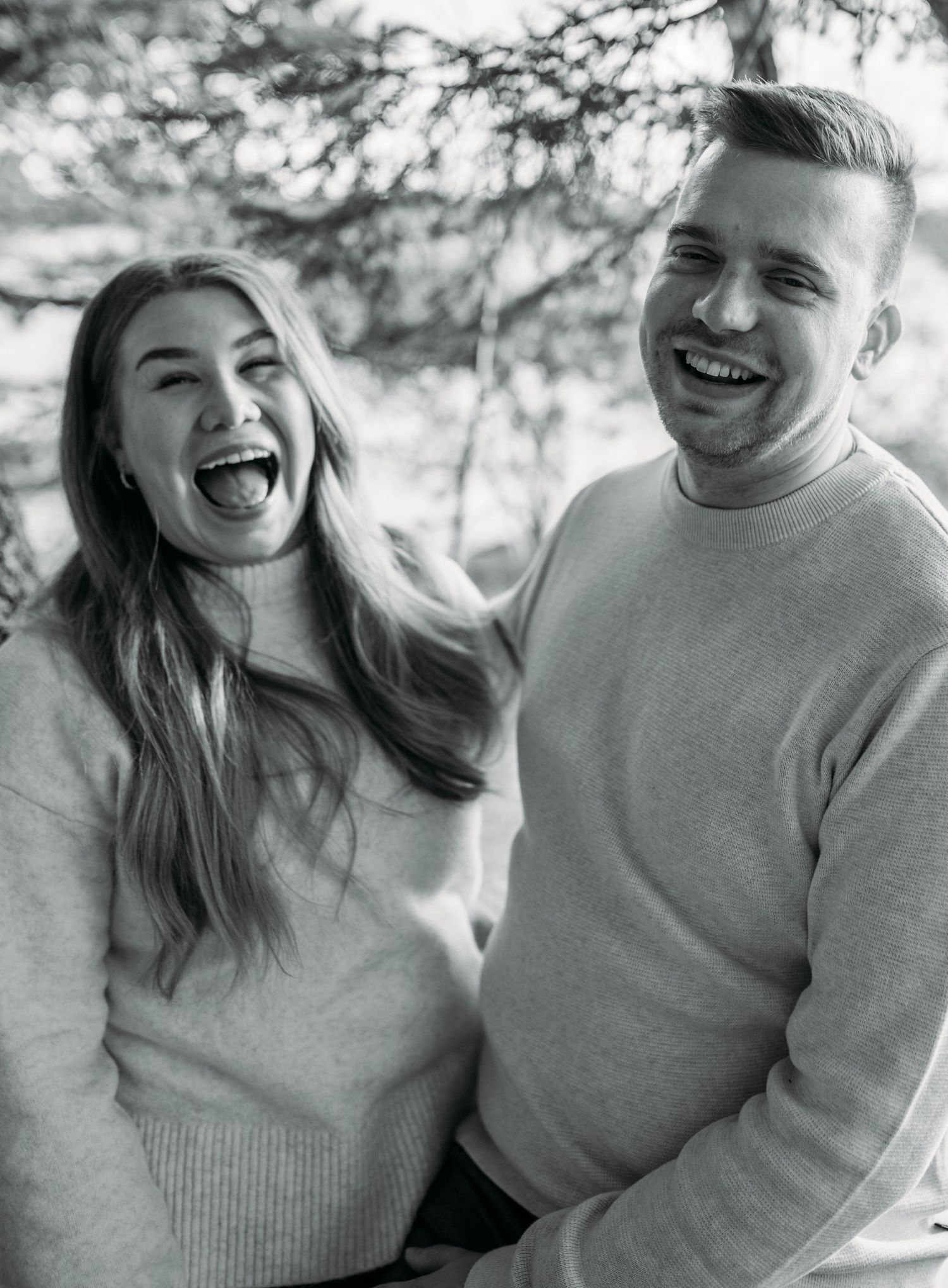 Black and white photo of a laughing couple in cozy sweaters outdoors, sharing a joyful moment together.