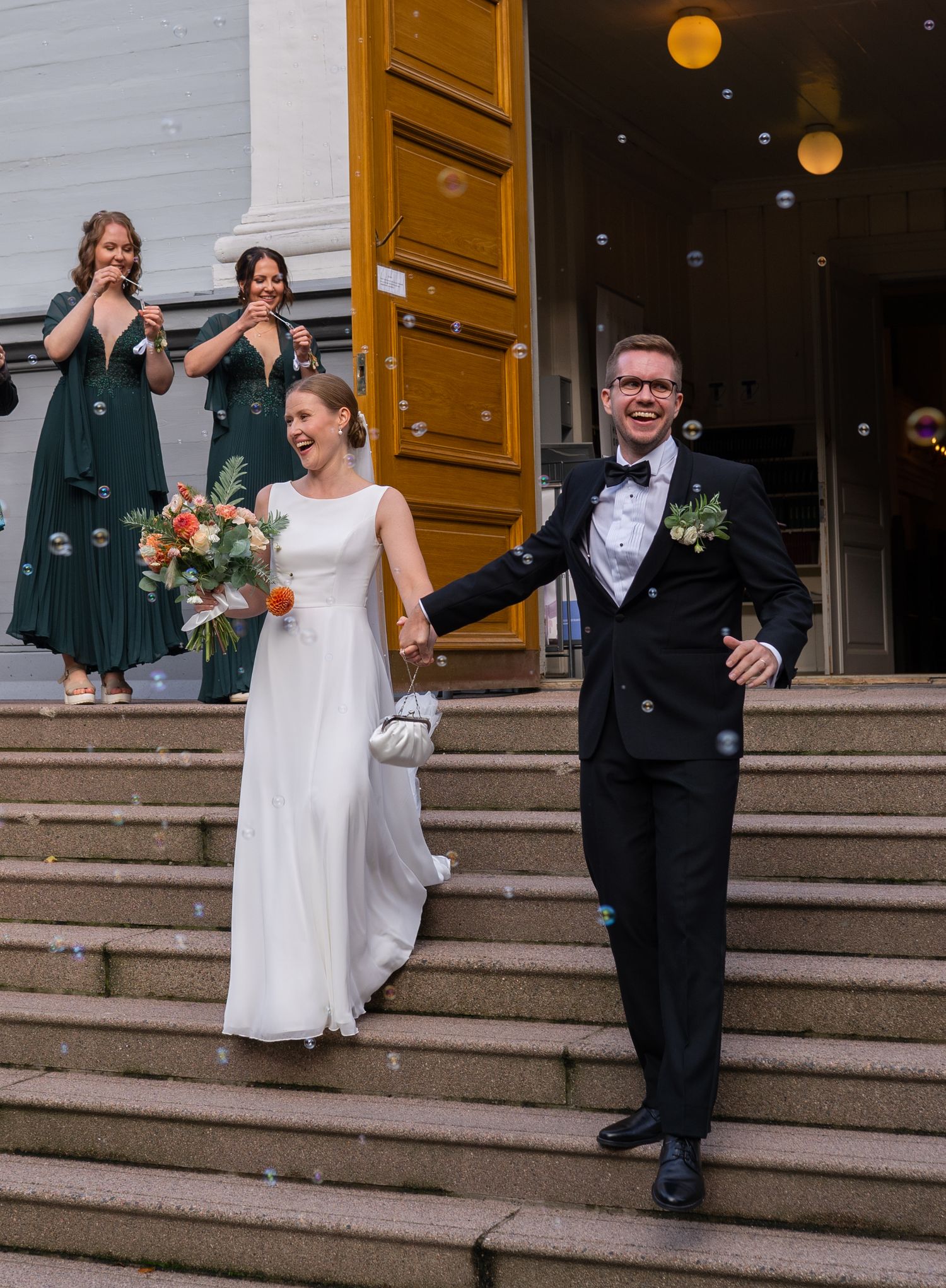 Joyful newlyweds descend church steps as bridesmaids blow bubbles in green dresses.