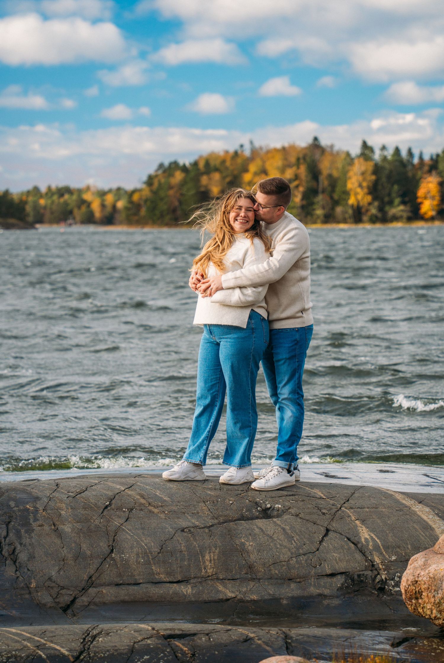 Couple in matching white sweaters and jeans embracing on rocks by a lake with autumn foliage in background.