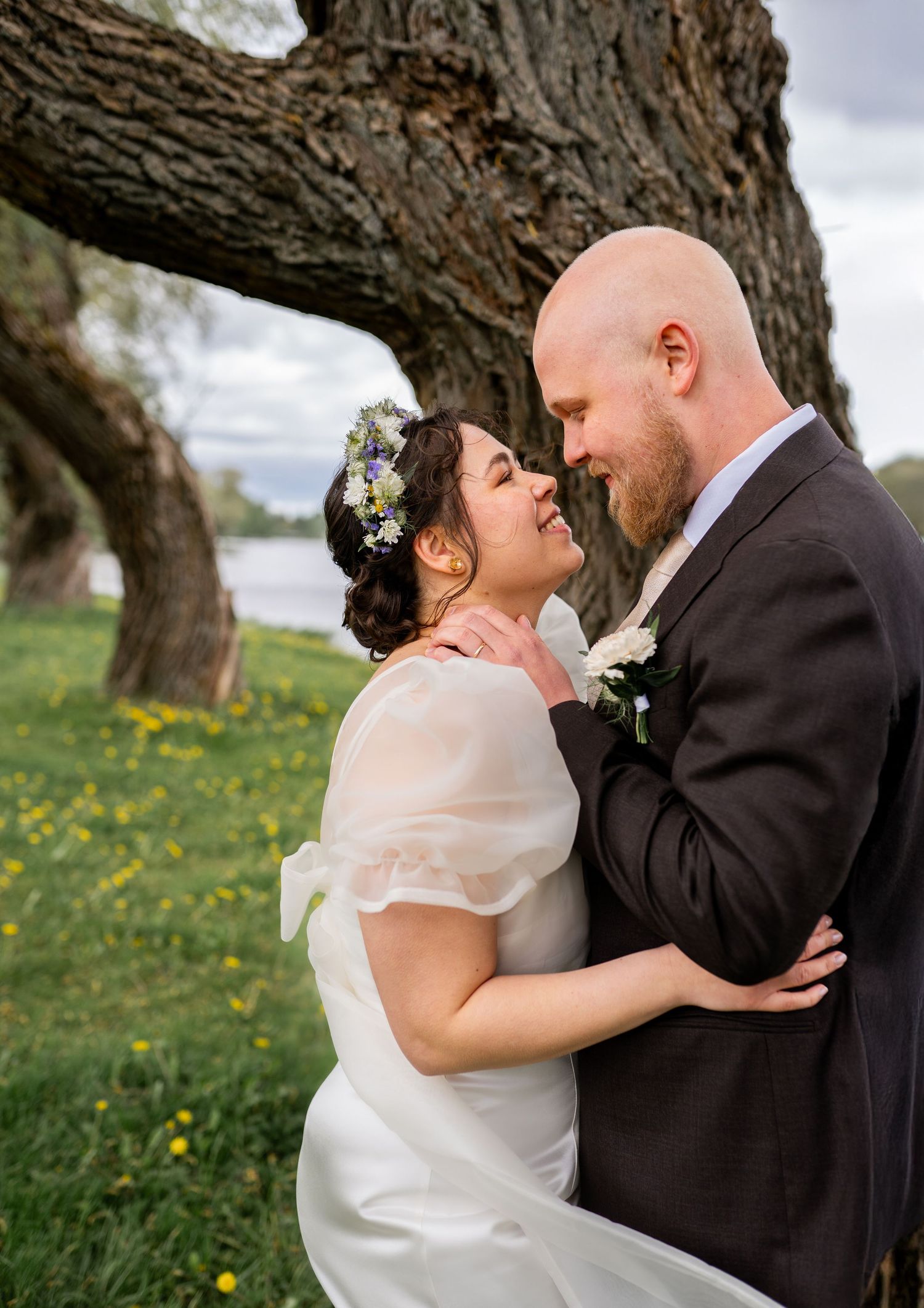 Bride in white dress with floral crown and groom in dark suit embracing under large tree with yellow wildflowers.