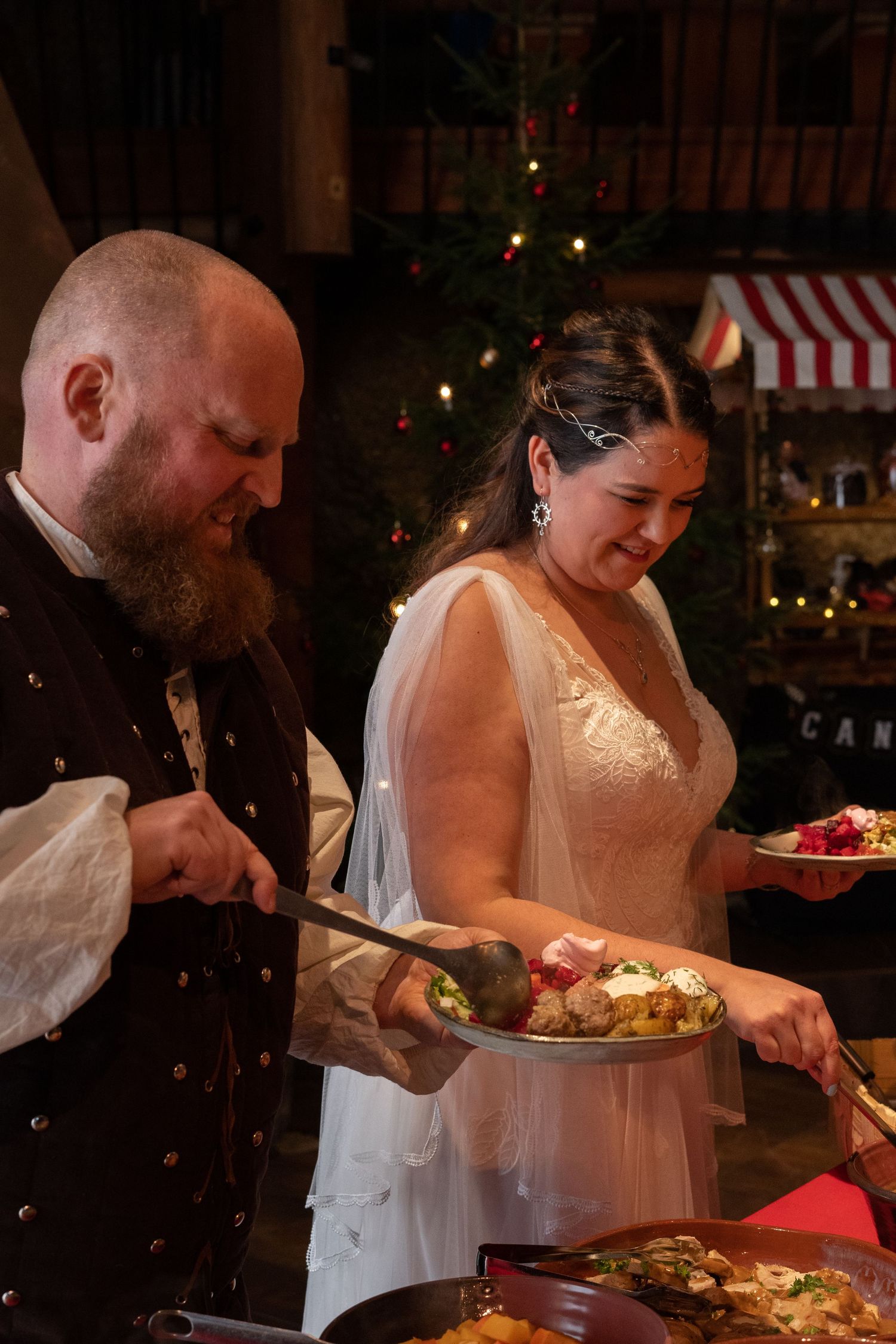 Bride and groom serving themselves at a wedding buffet dinner, with a decorated Christmas tree in the background.