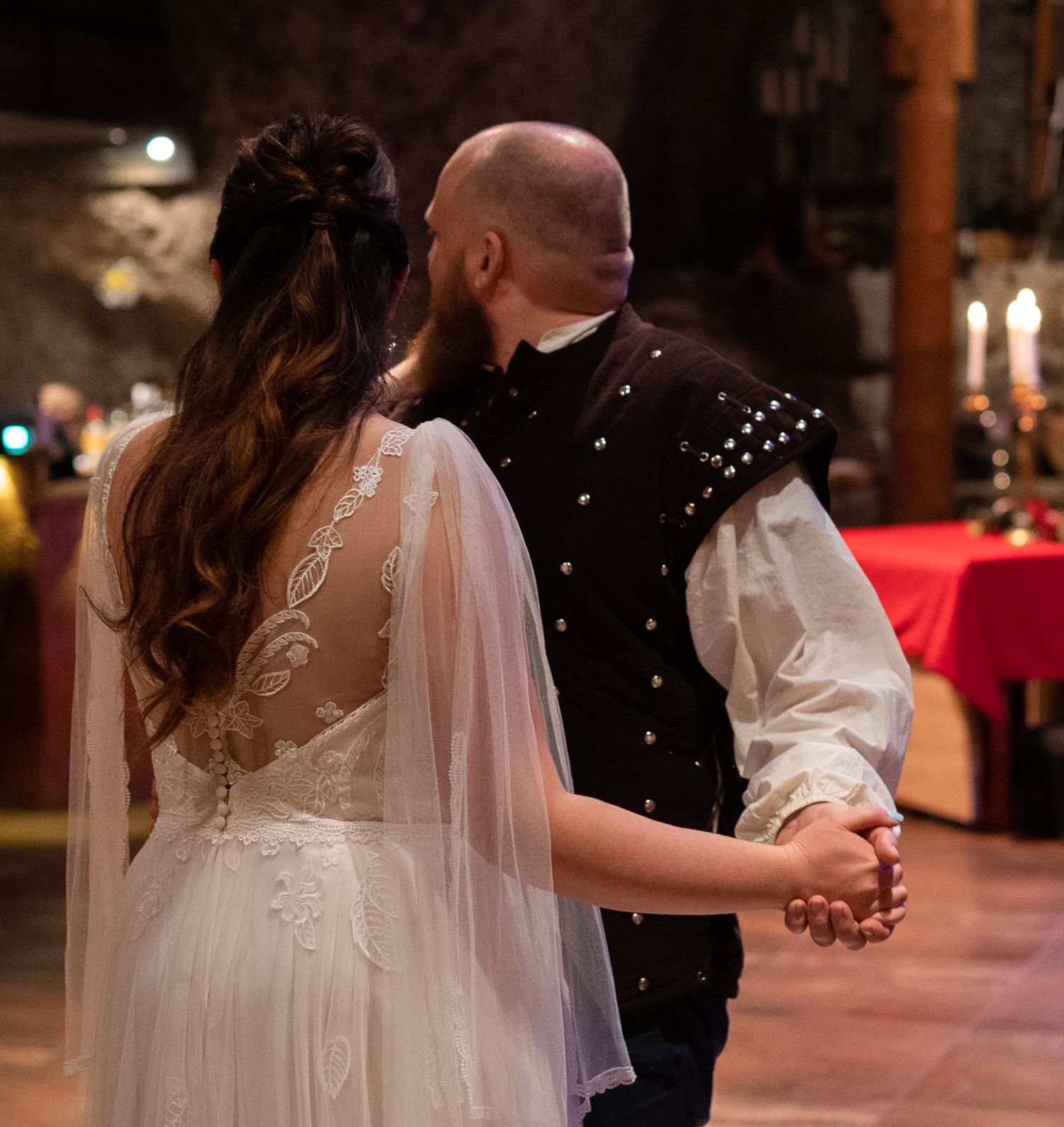 A bride and groom share a first dance in a candlelit stone hall, wearing elegant medieval-inspired wedding attire.