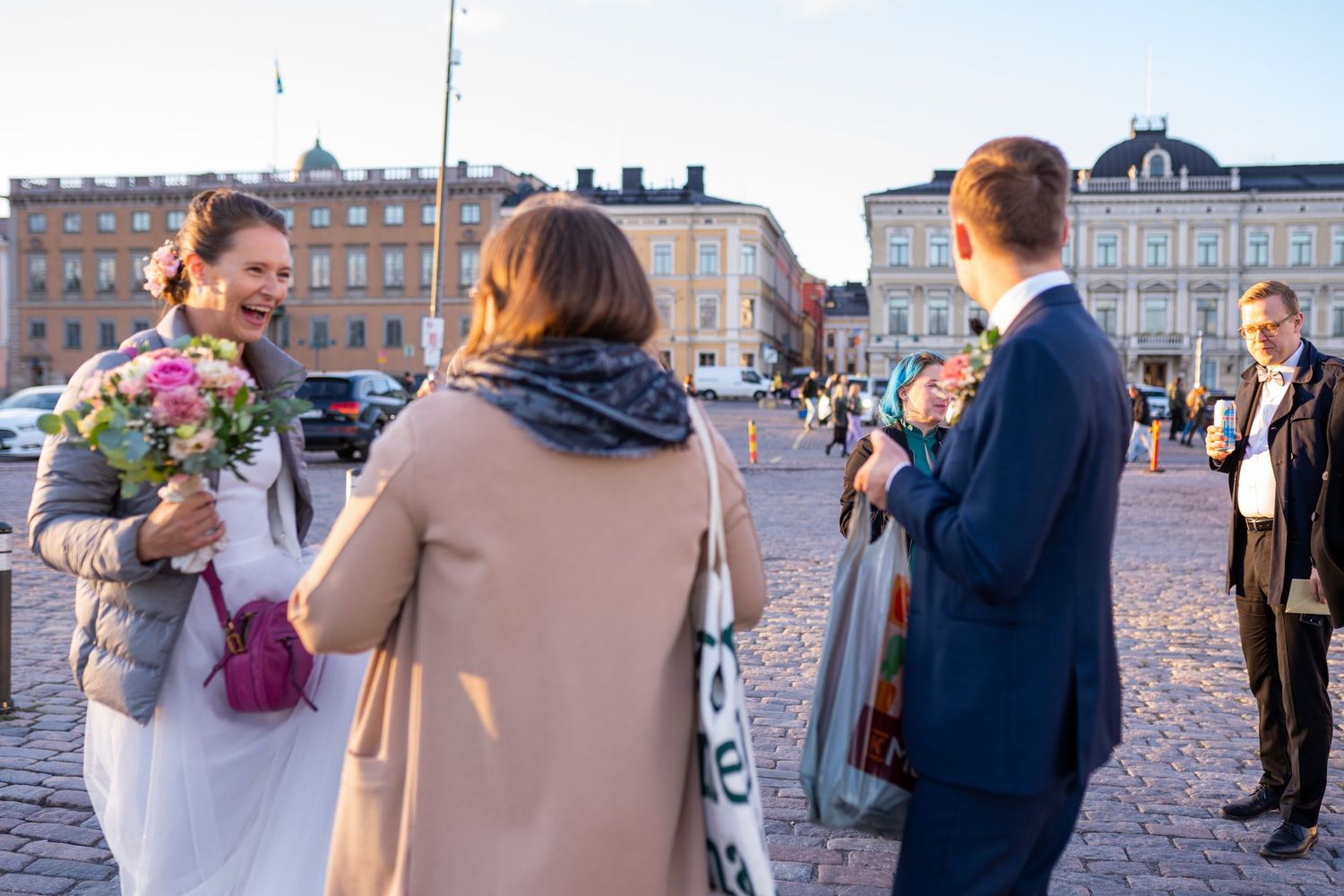 Bride holding bouquet greets guests in a sunny city square.