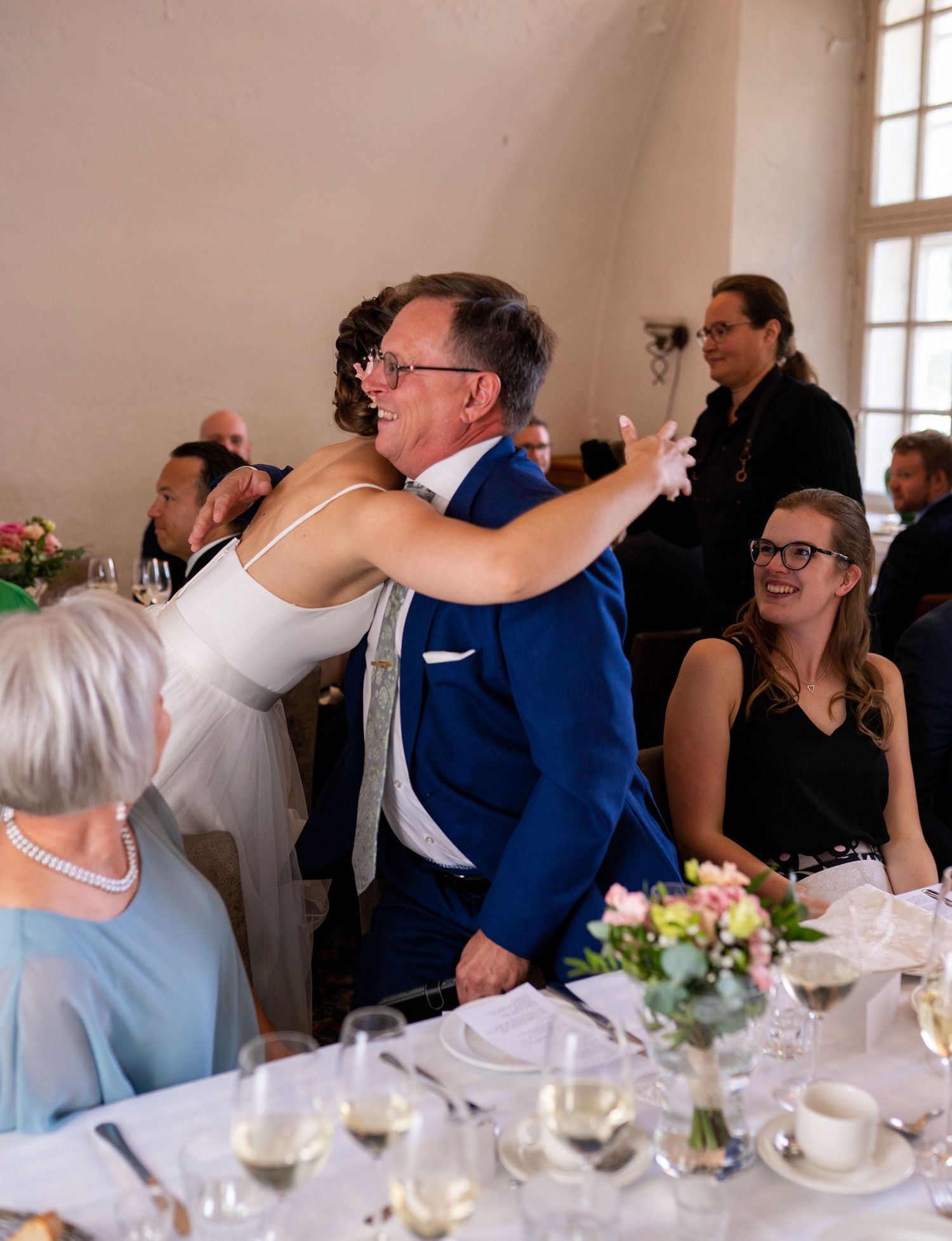 Bride in white dress embracing man in blue suit at wedding reception table with smiling guests.