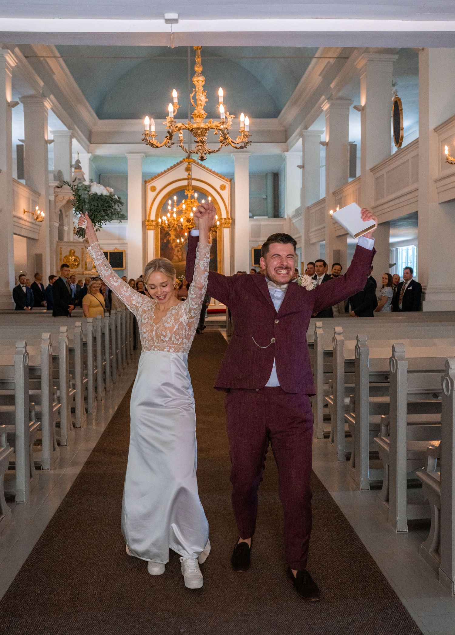 Joyful newlyweds raise hands in celebration while walking down church aisle after wedding ceremony.