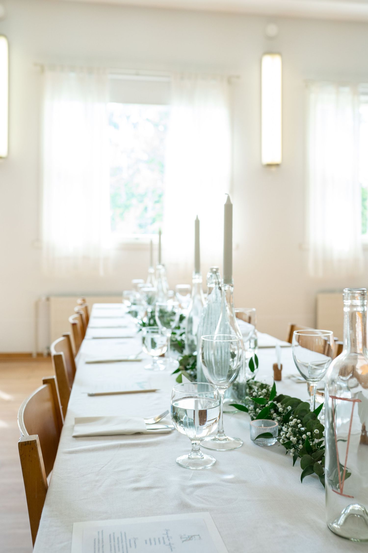 Elegant wedding reception table set with white linen, candles, glassware, and greenery garland centerpiece.