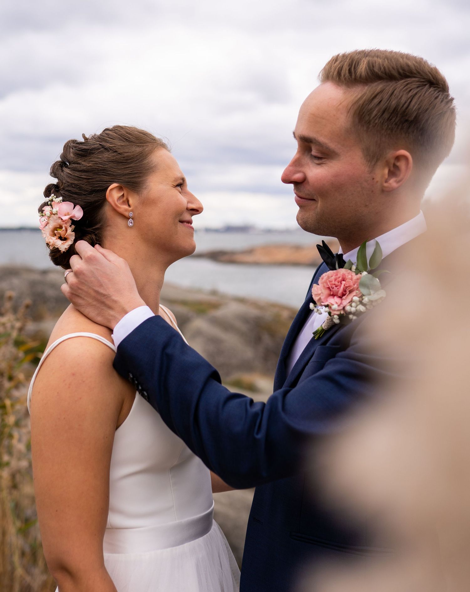 Bride and groom gazing at each other, she wears floral hair accessory, he has pink boutonniere, coastal backdrop.