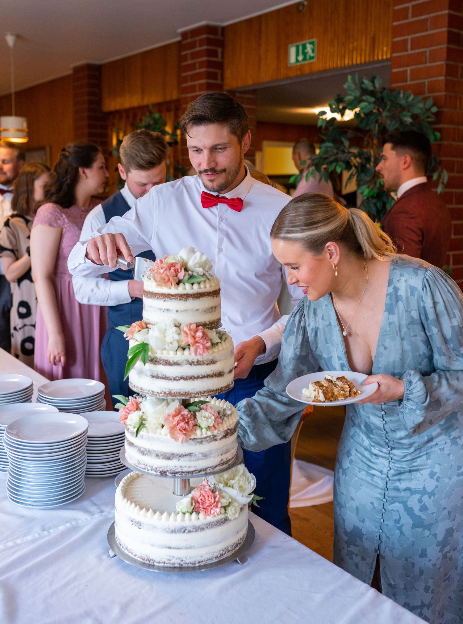 Couple cutting a three-tier naked wedding cake with pink floral decorations at a wedding reception.