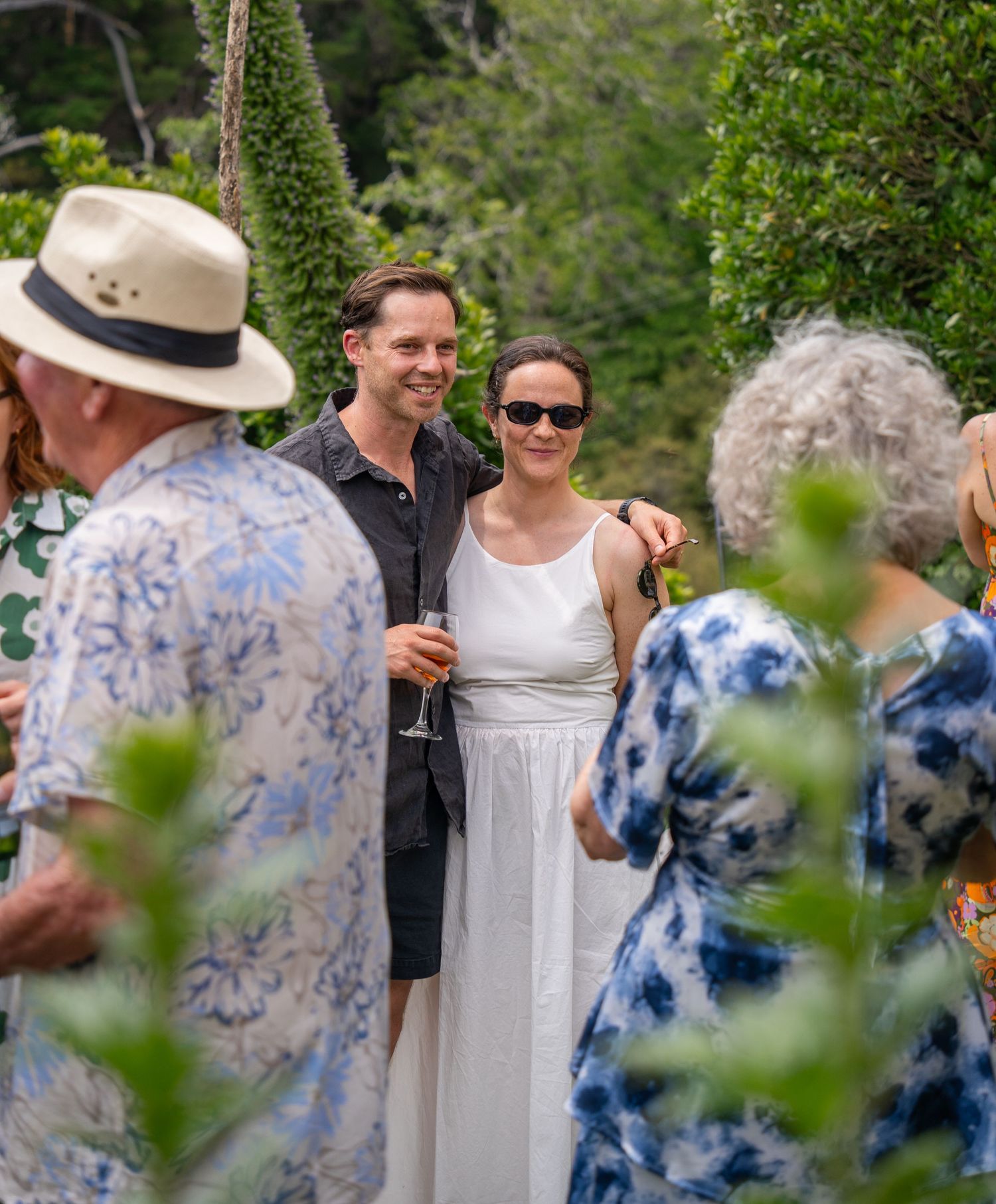Couple smiling together at an outdoor garden party surrounded by guests in summer attire.