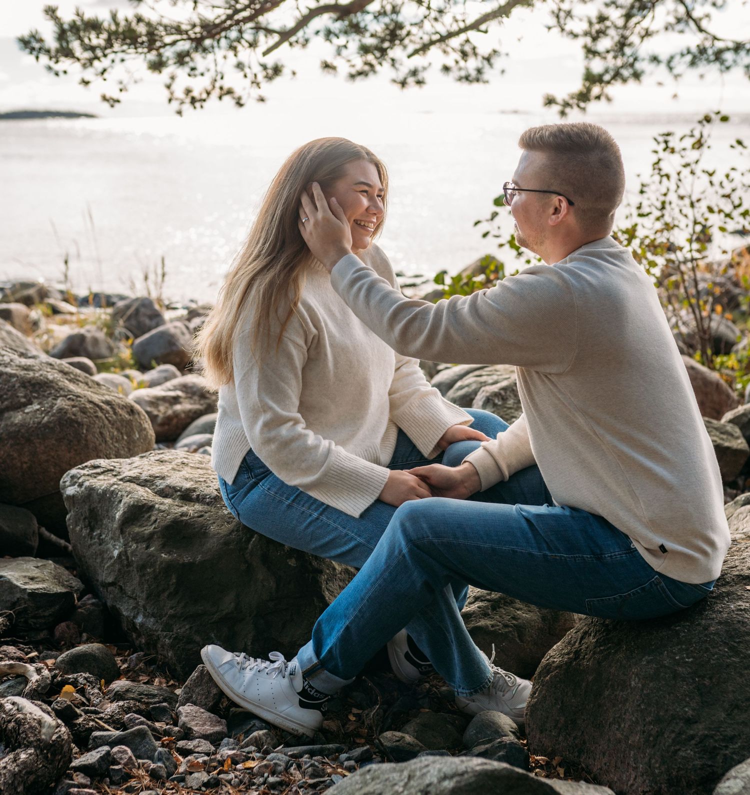 Couple sitting on rocks by the water, man gently touching woman's hair, both smiling and wearing jeans.