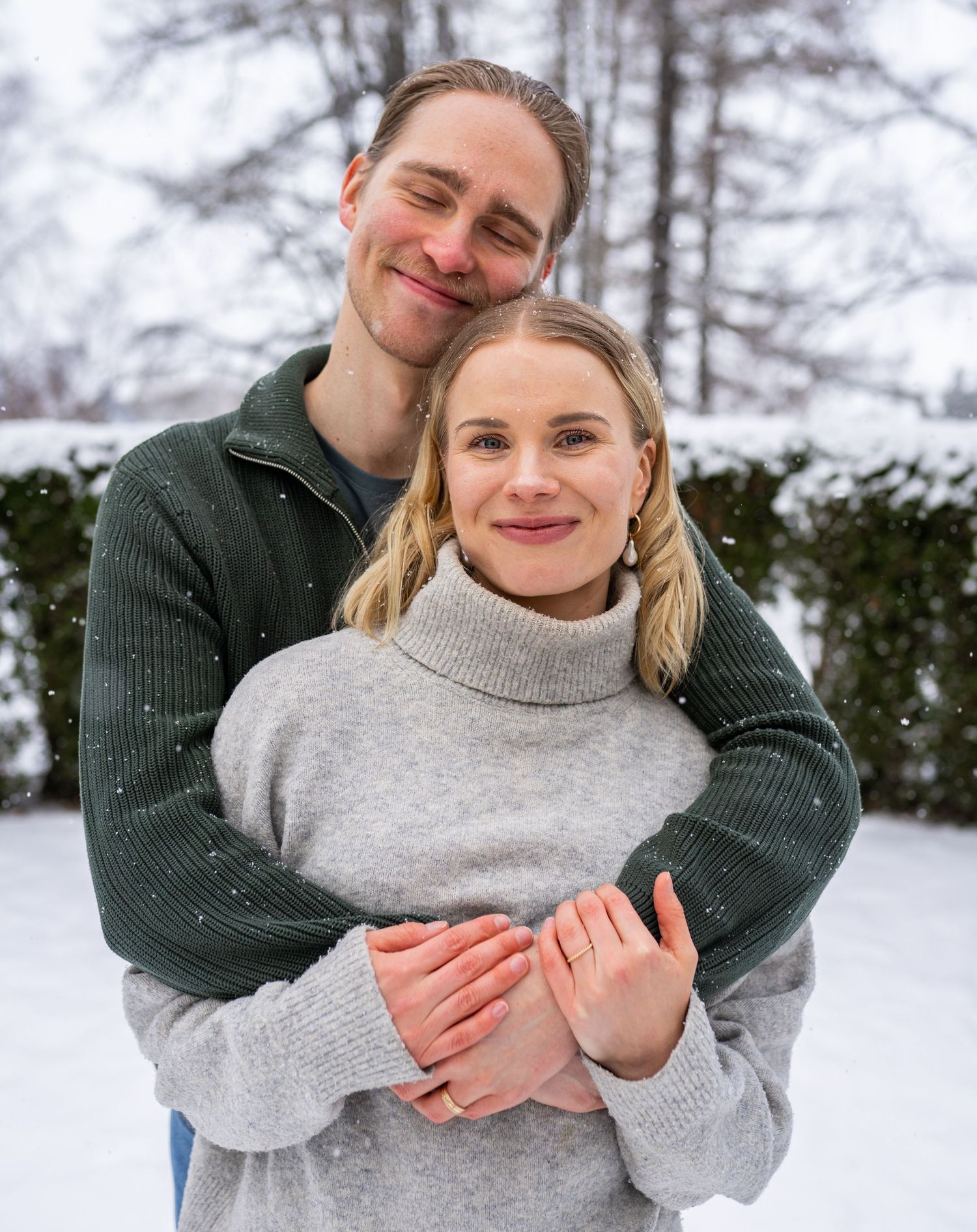 A couple embraces warmly in a snowy winter setting, wearing cozy sweaters, smiling happily outdoors.