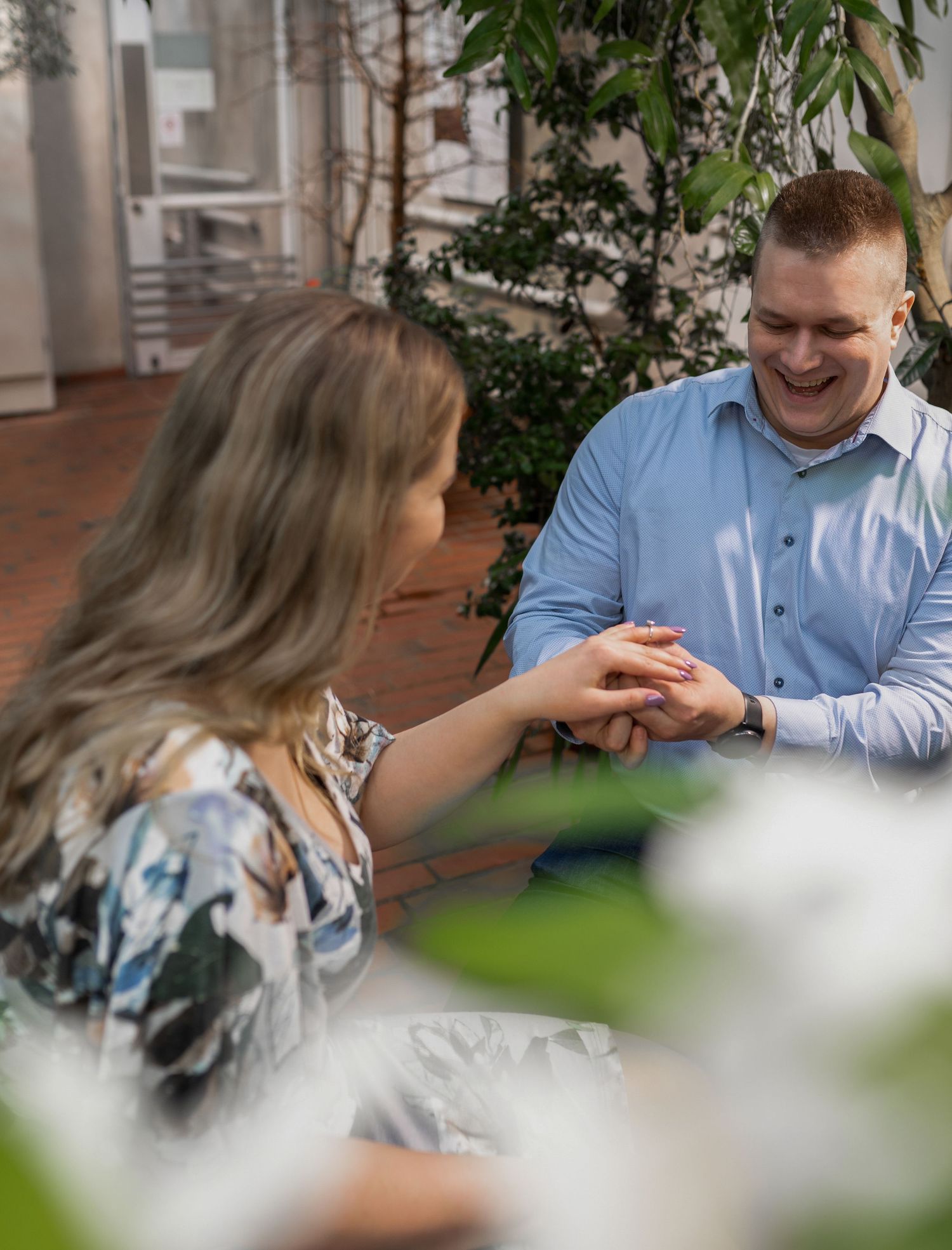 A smiling man in a blue shirt places an engagement ring on a woman's finger at an outdoor restaurant.