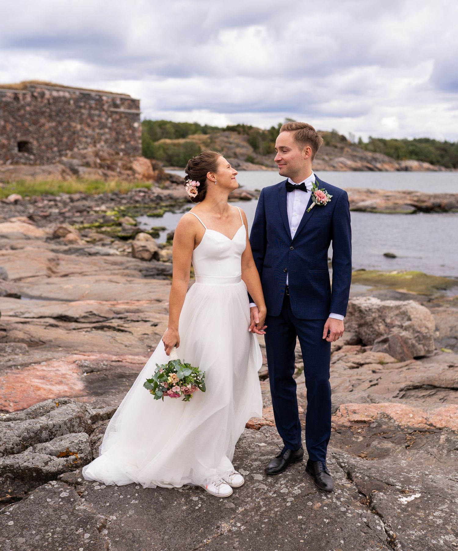 Wedding couple holding hands on rocky shore, bride in white gown with bouquet, groom in navy suit, coastal backdrop.