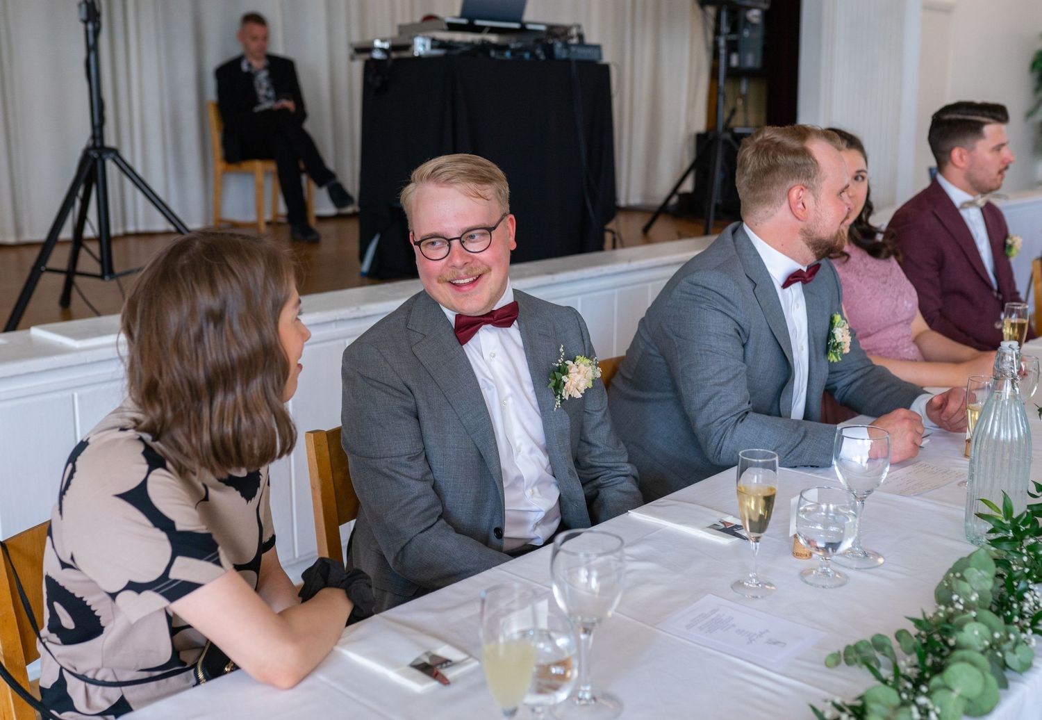 Smiling wedding guests seated at decorated reception table with DJ in background.