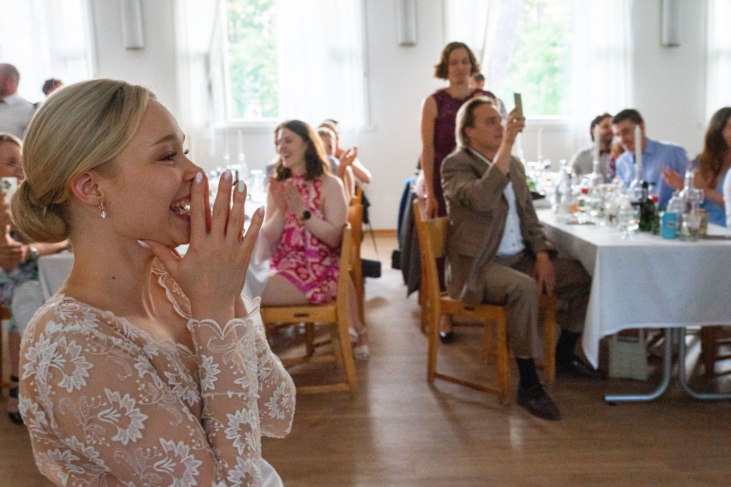 Delighted bride laughing and covering mouth while guests clap during wedding reception surprise.
