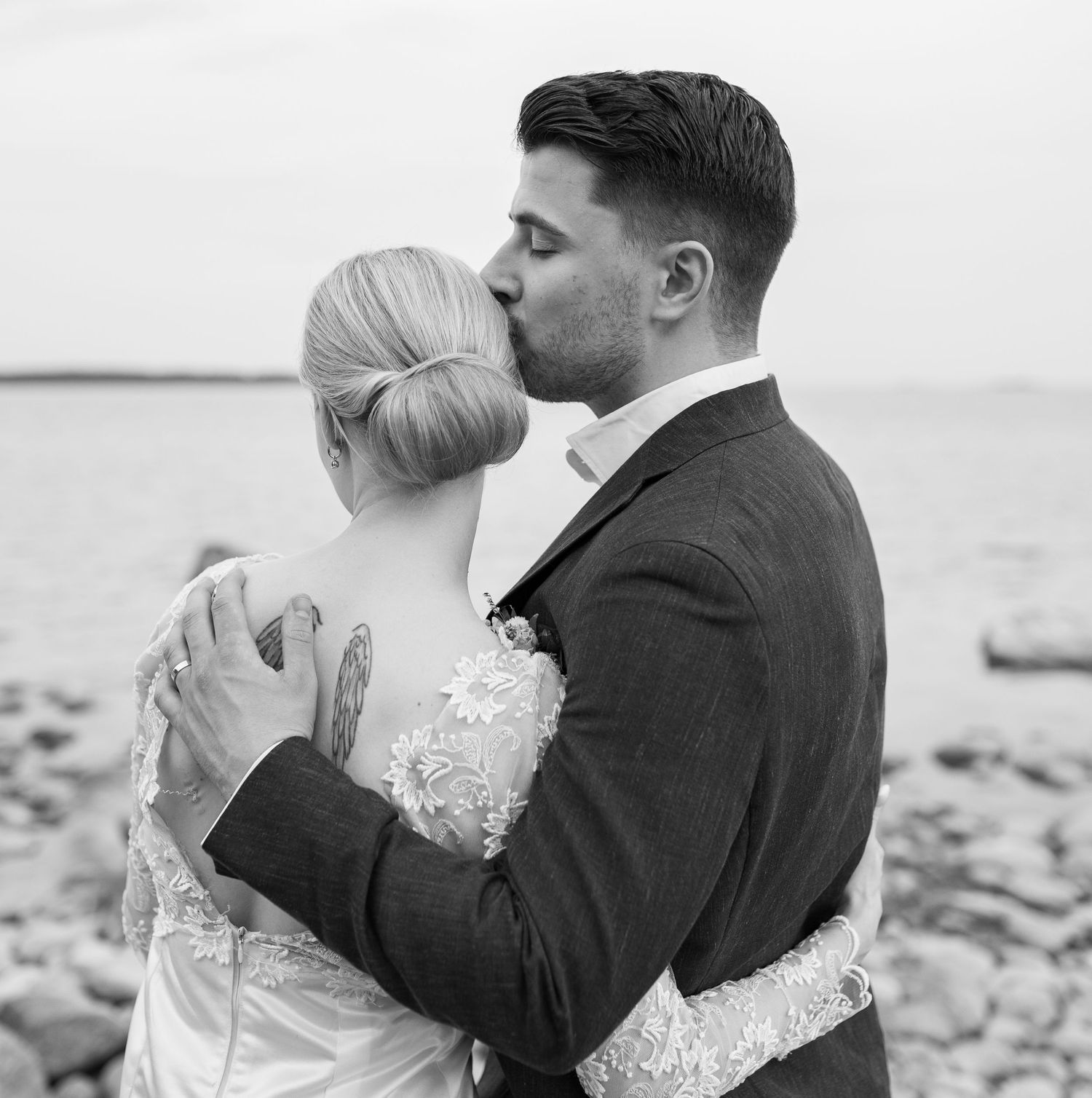 Black and white photo of groom kissing bride on cheek while embracing on a rocky seaside beach.